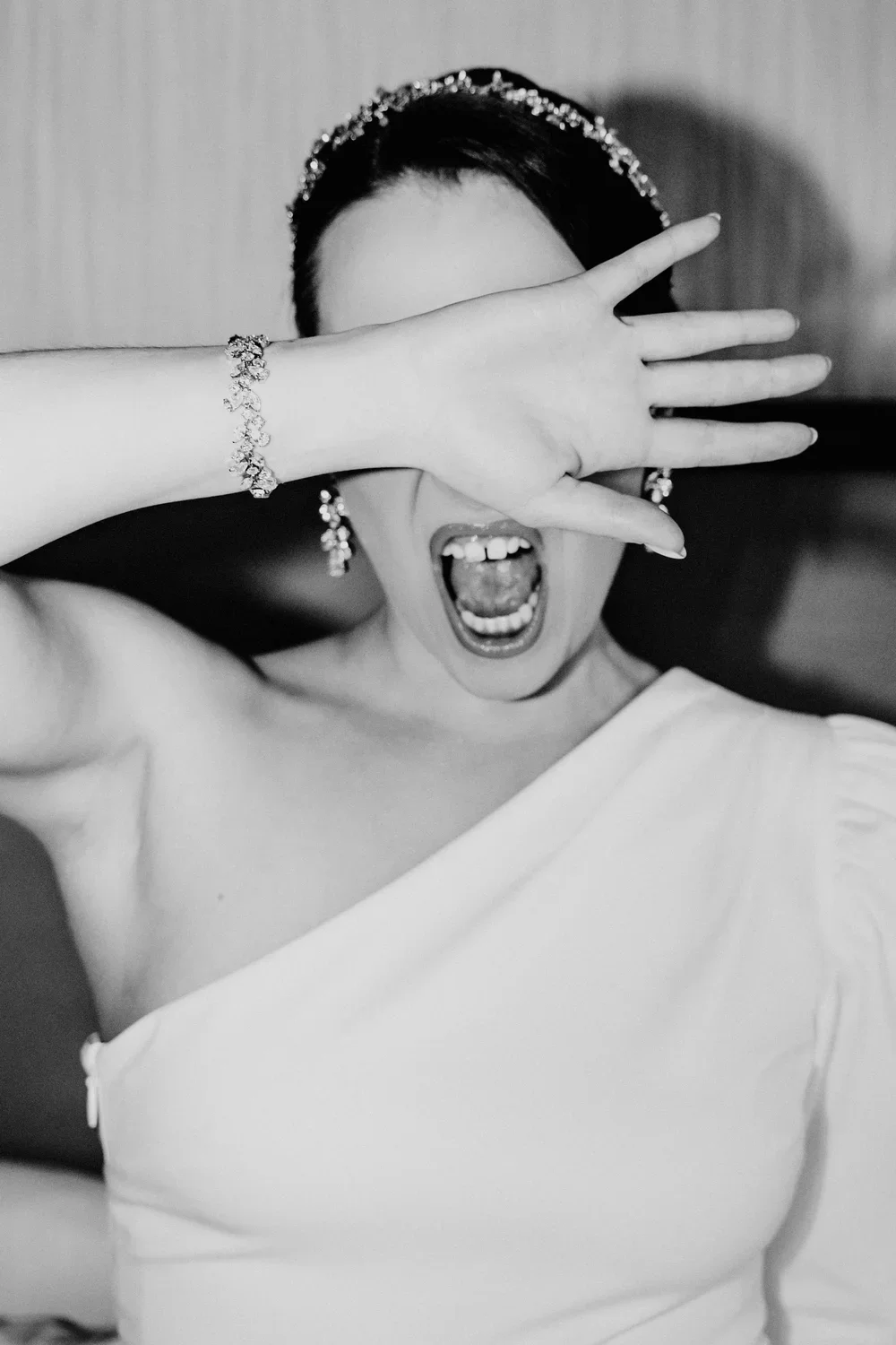 A woman in a white dress with jewelry on her wrist and earrings, covering her face with her hand and shouting.