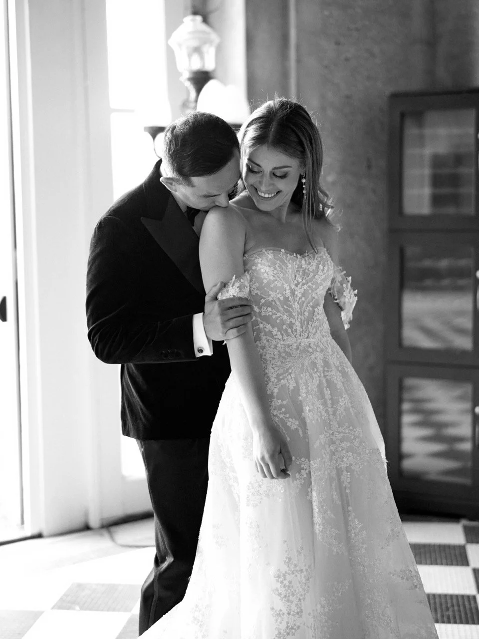 A groom in a dark tuxedo is helping a bride in a wedding dress adjust her gown and they are smiling. The scene is indoors with a window and a cabinet in the background.