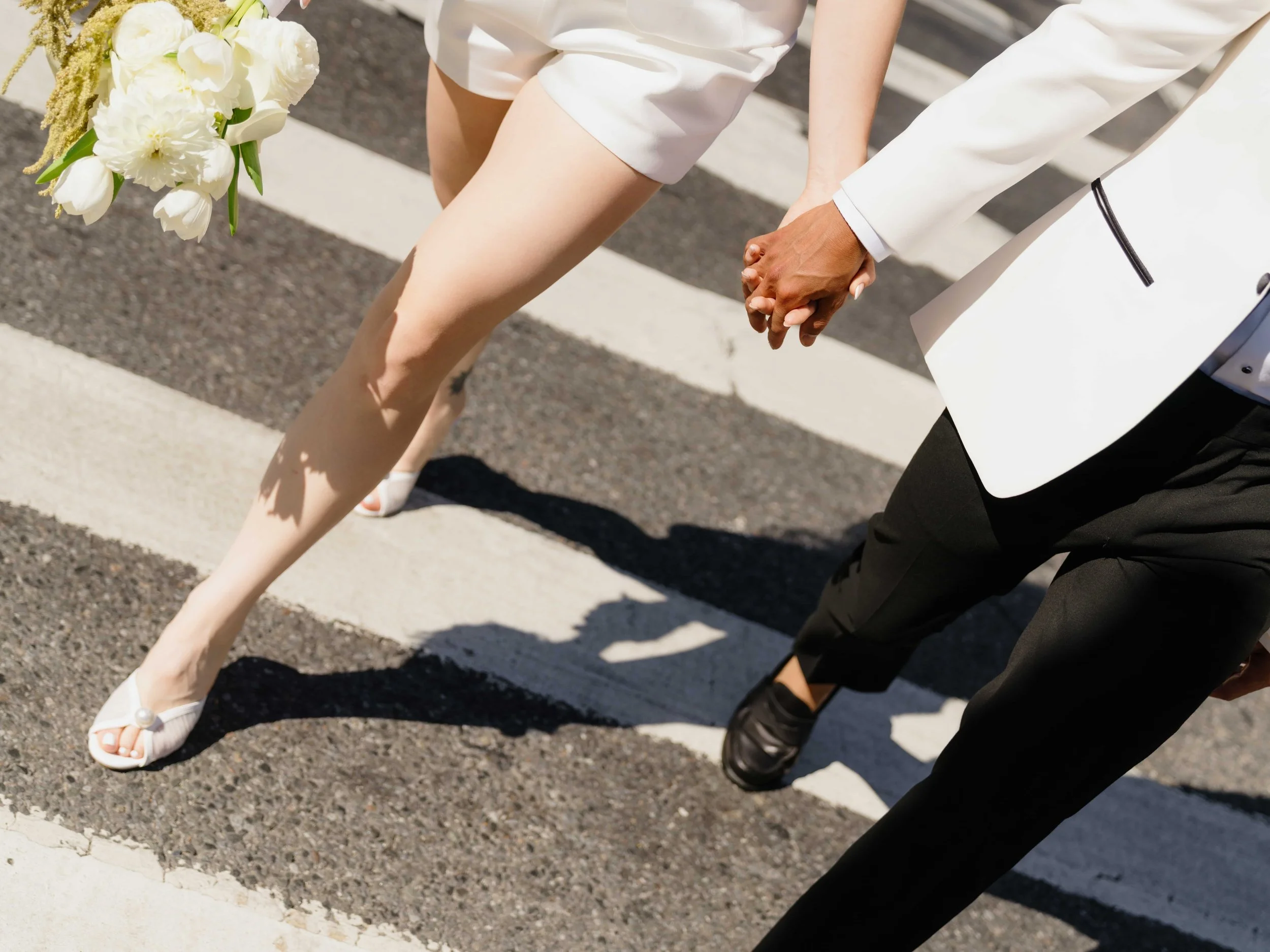 A couple holding hands while walking across a crosswalk. The woman is wearing a white dress and high heels, holding a bouquet of flowers. The man is wearing black pants and a white blazer.