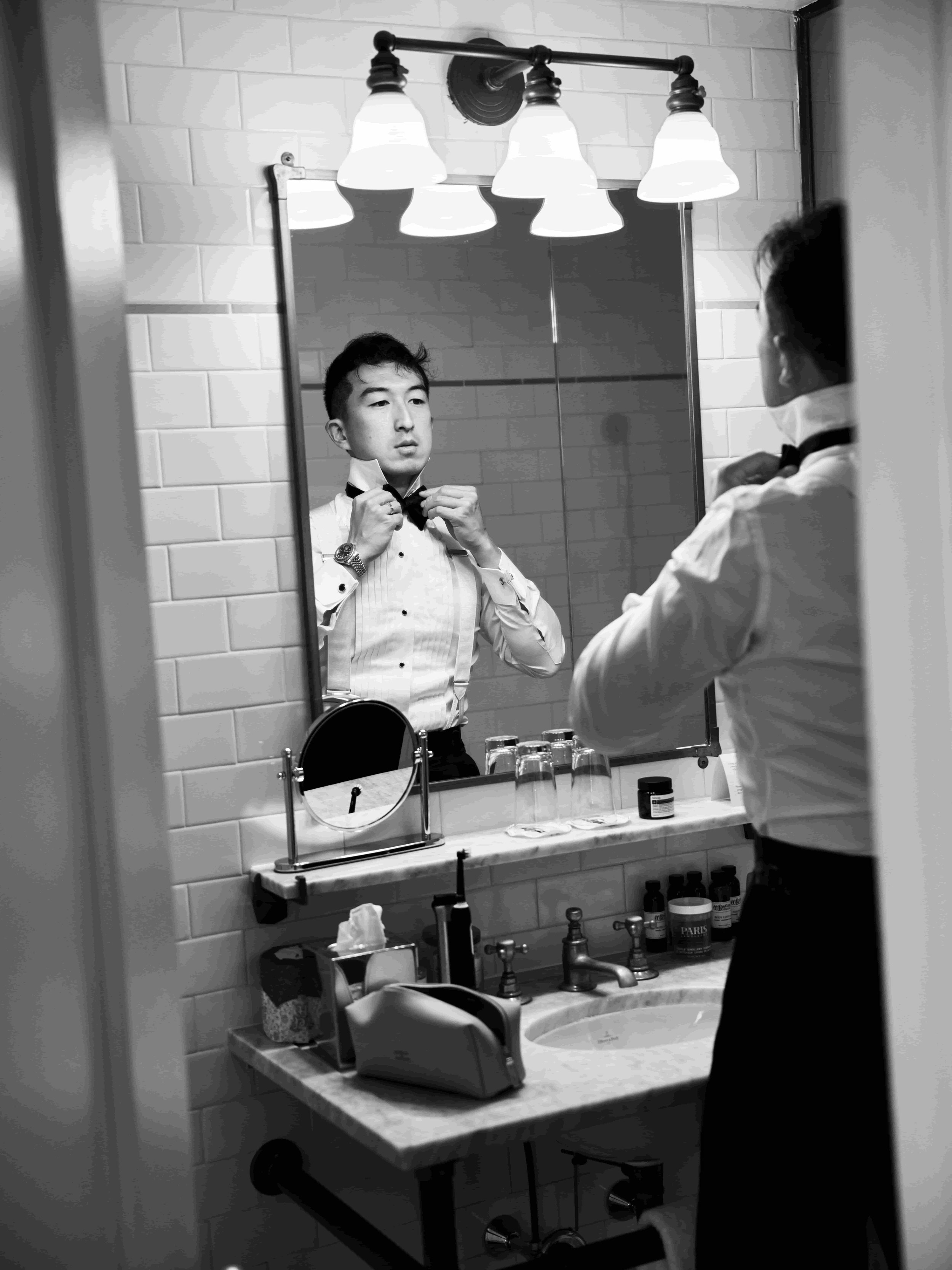 A man in a tuxedo adjusting his bowtie while looking at his reflection in a bathroom mirror. The bathroom has white subway tiles, a marble countertop, and various grooming items on the counter, with a vanity mirror and a light fixture above.