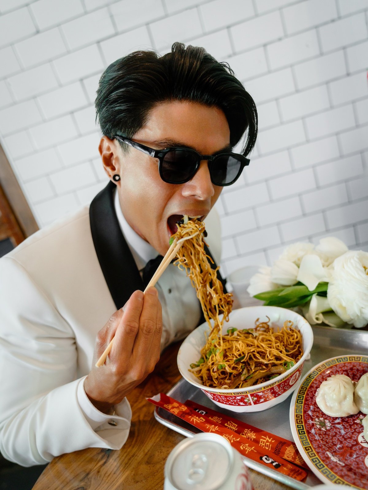 A man with dark hair, sunglasses, and a tuxedo shirt is eating noodles with chopsticks at a table with traditional Chinese dishes, including a bowl of noodles and dumplings.