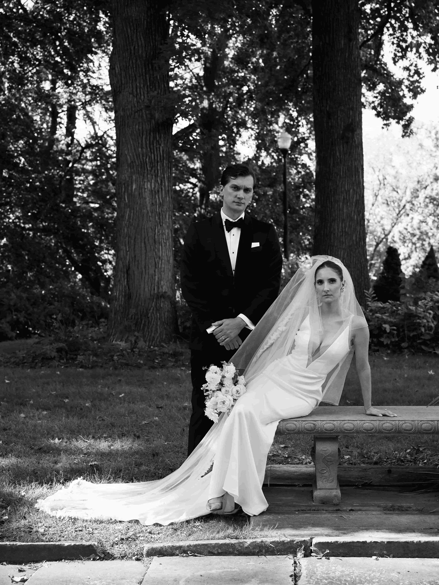 Black and white photo of a bride in a white wedding dress with a veil, sitting on a park bench holding a bouquet of flowers, and a groom in a tuxedo standing next to her, outdoors with trees in the background.