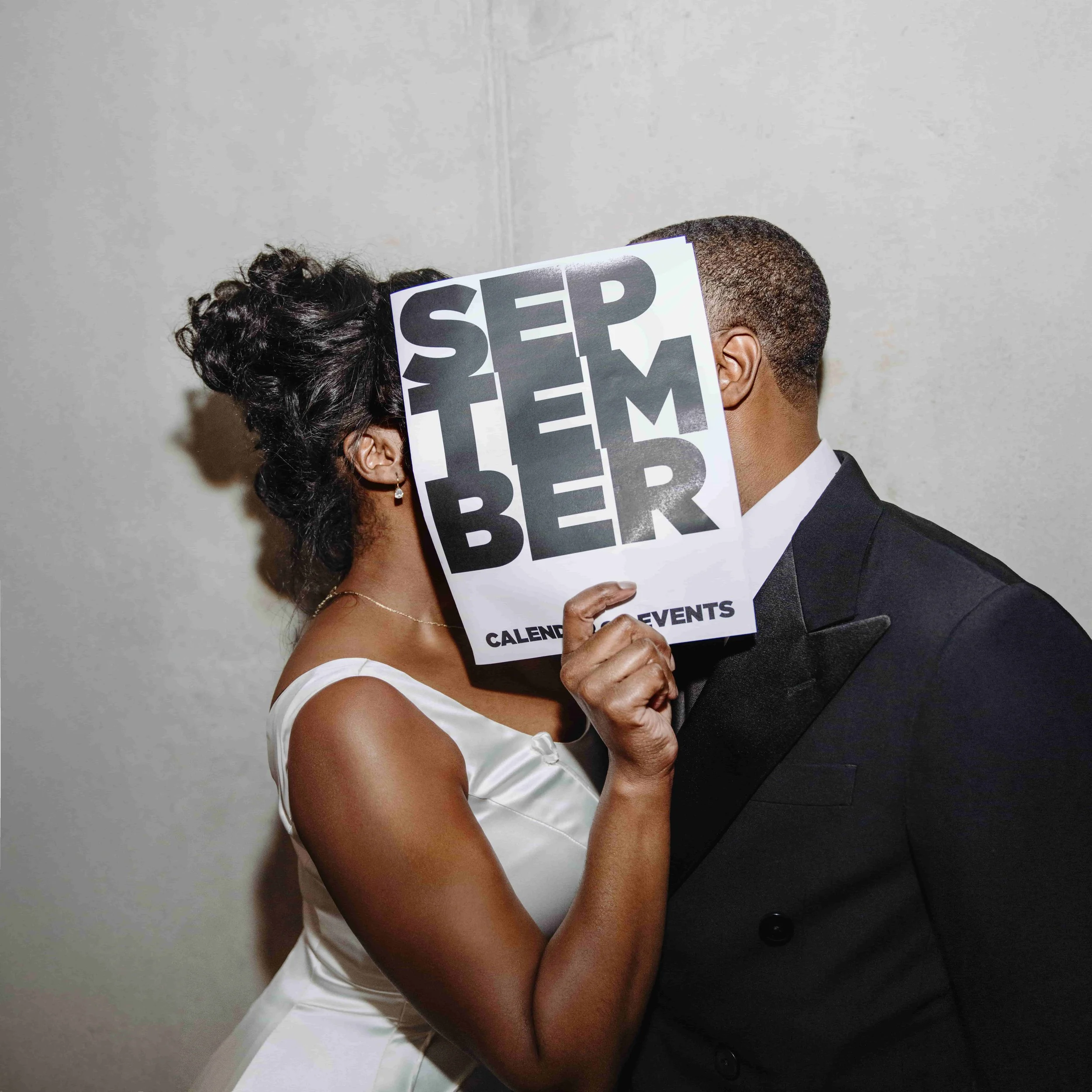 A woman and a man are standing close, with the woman holding a sign that covers part of their faces. The woman is wearing a white dress and has dark, curly hair. The man is dressed in a black tuxedo. The sign says 'SEPTEMBER' in large black letters, 