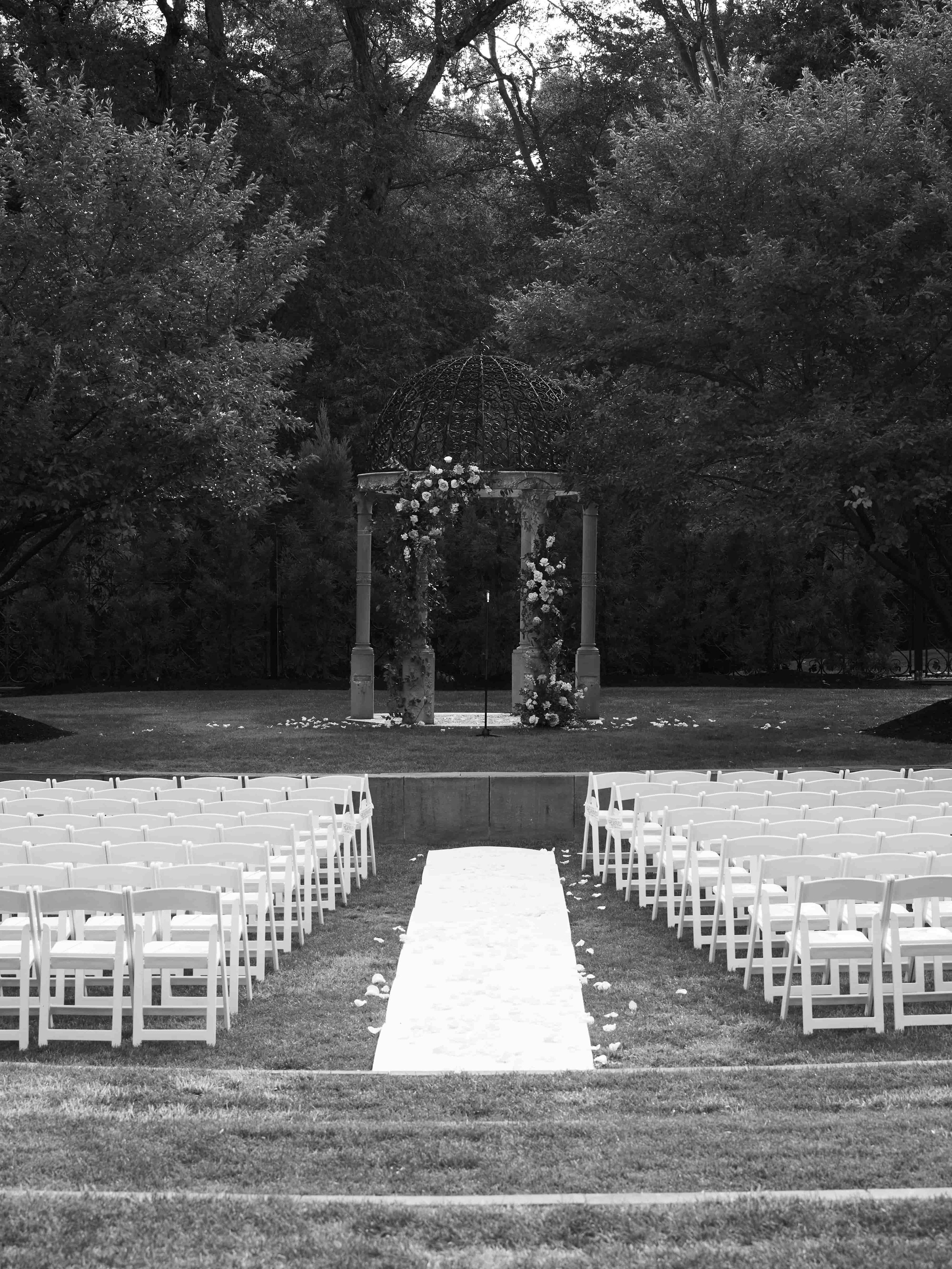 Black and white photo of an outdoor wedding ceremony setup with chairs arranged on both sides of an aisle, leading to a decorated gazebo in a garden surrounded by trees.