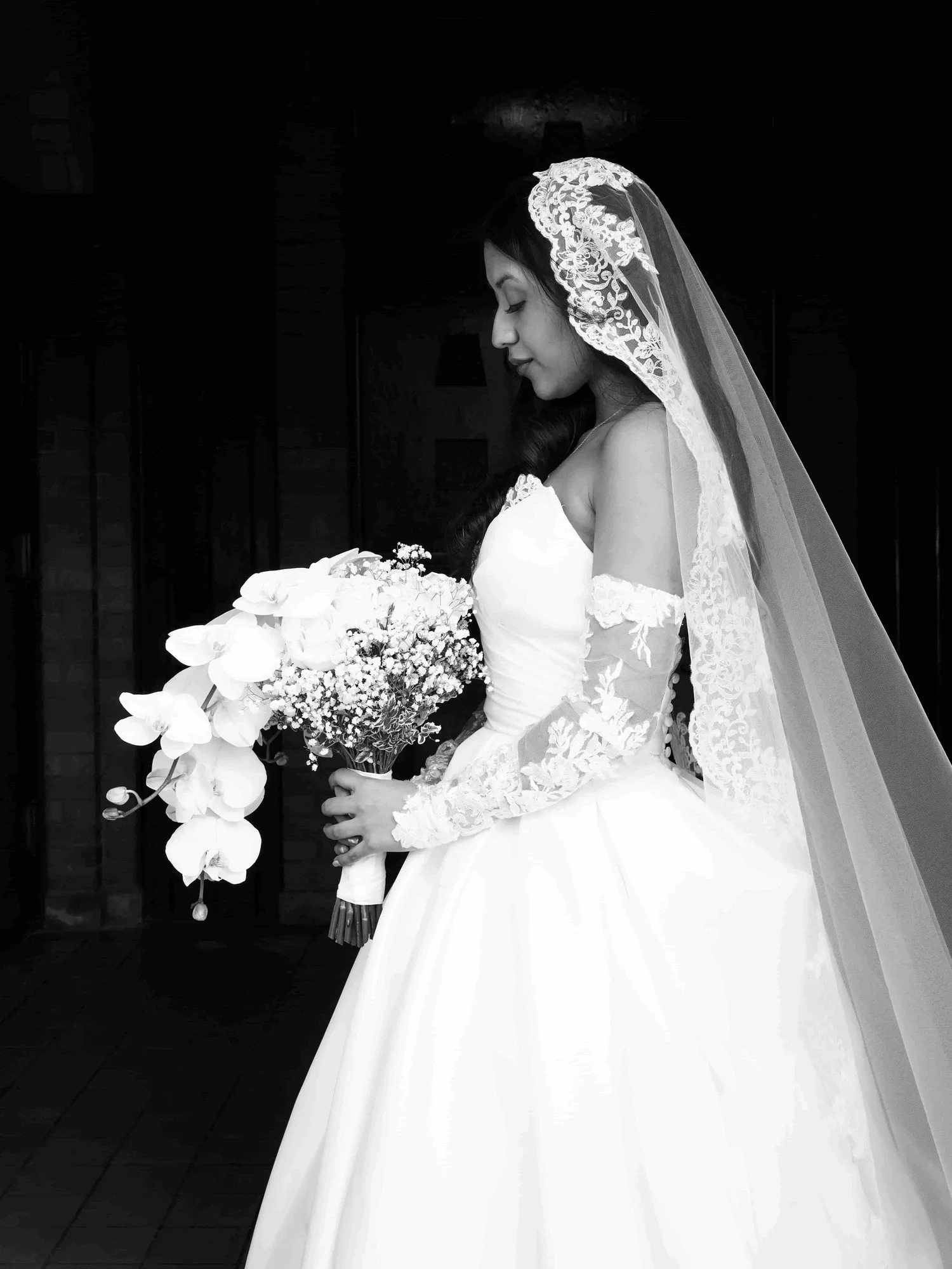 Black and white photo of a bride holding a bouquet of flowers, dressed in a wedding gown with lace details and a veil, standing in profile against a dark background.