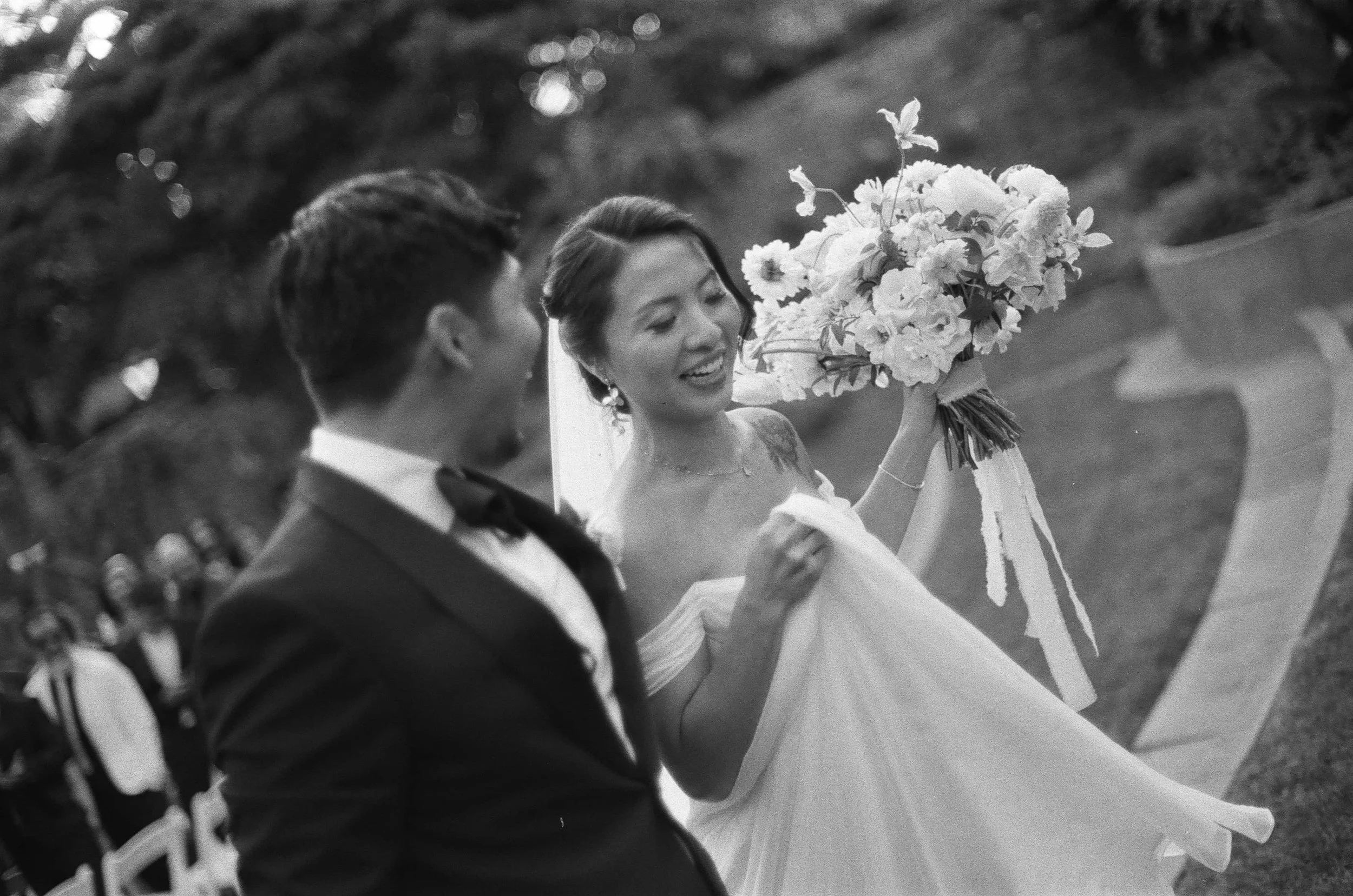 A bride holding a bouquet of flowers and smiling at a groom at an outdoor wedding ceremony.