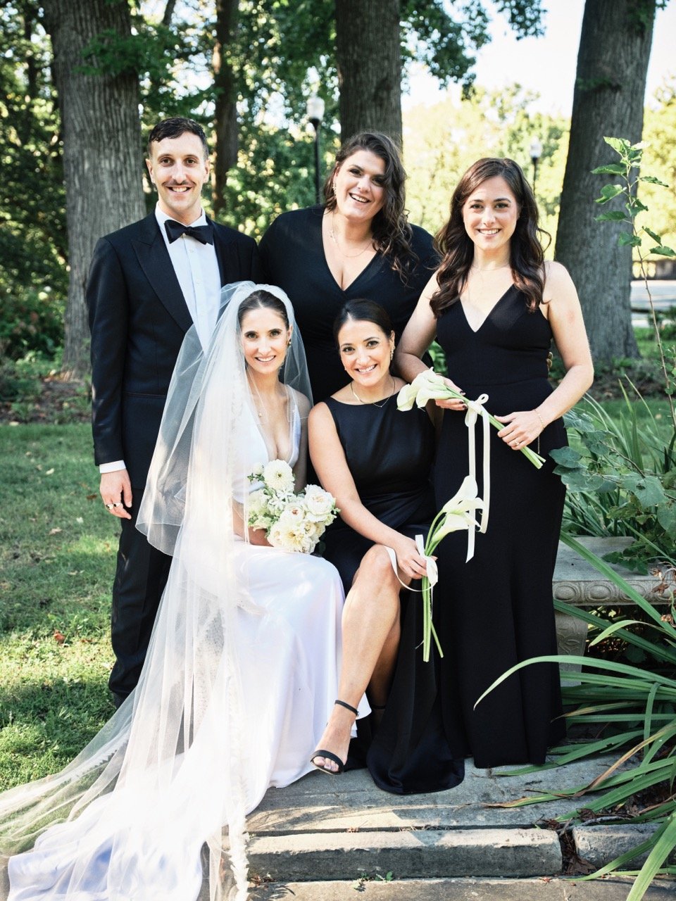 A bride in a white wedding dress and veil holds a bouquet, surrounded by four women in black dresses and a man in a tuxedo at an outdoor wedding with trees in the background.