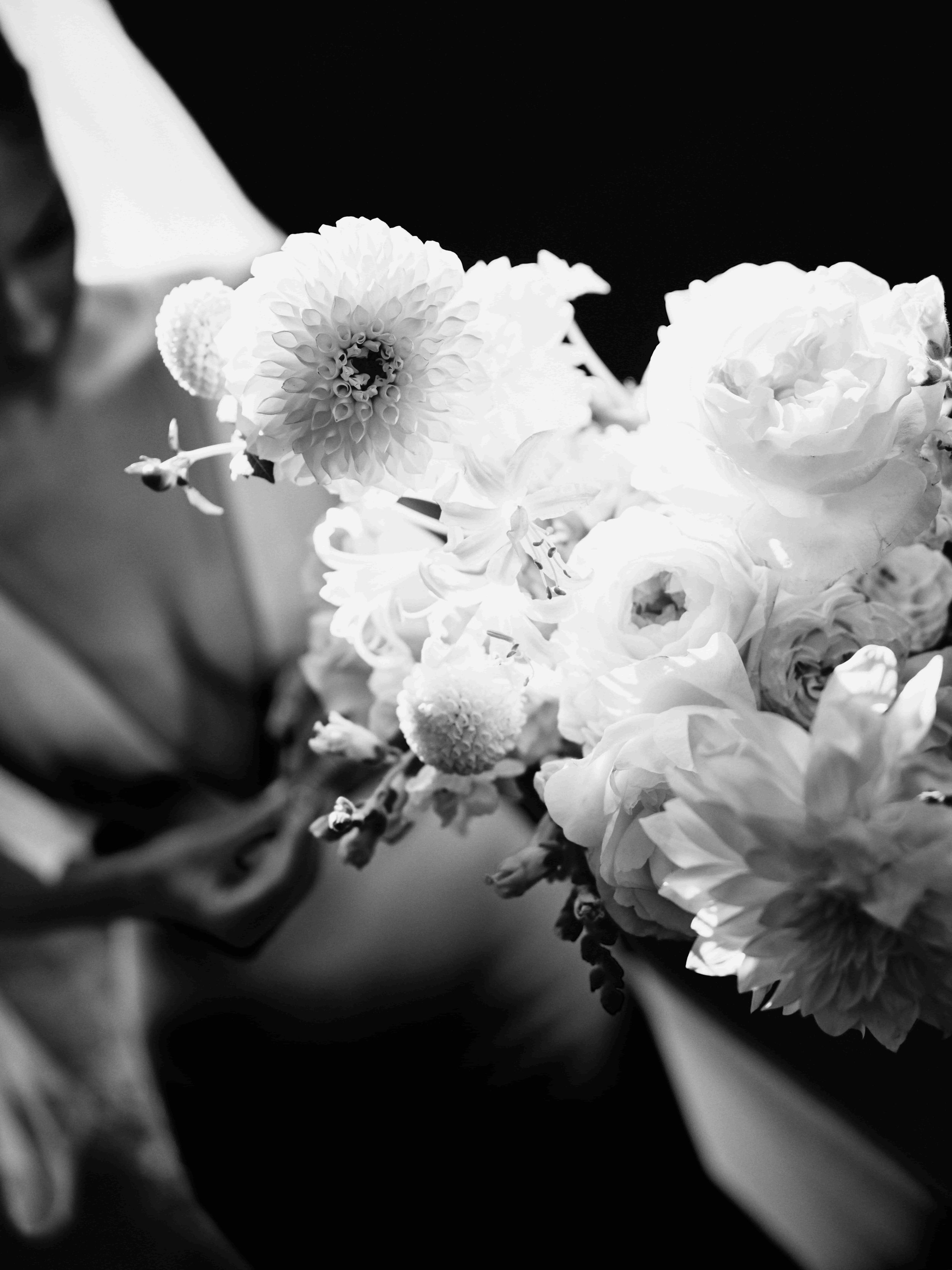 Black and white close-up of a bouquet of various flowers, including dahlias and roses, held by someone wearing a suit.
