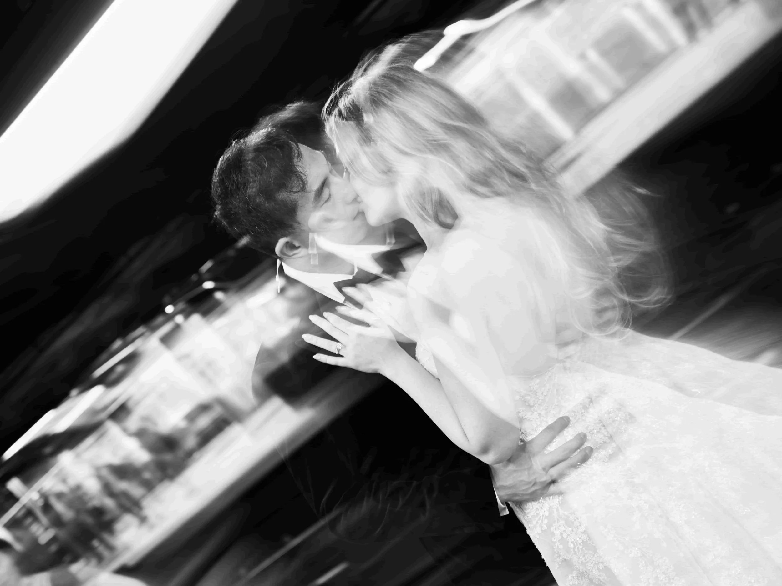 Black and white photograph of a newlywed couple sharing a kiss with their foreheads touching, the bride wearing a wedding dress and the groom in a tuxedo.