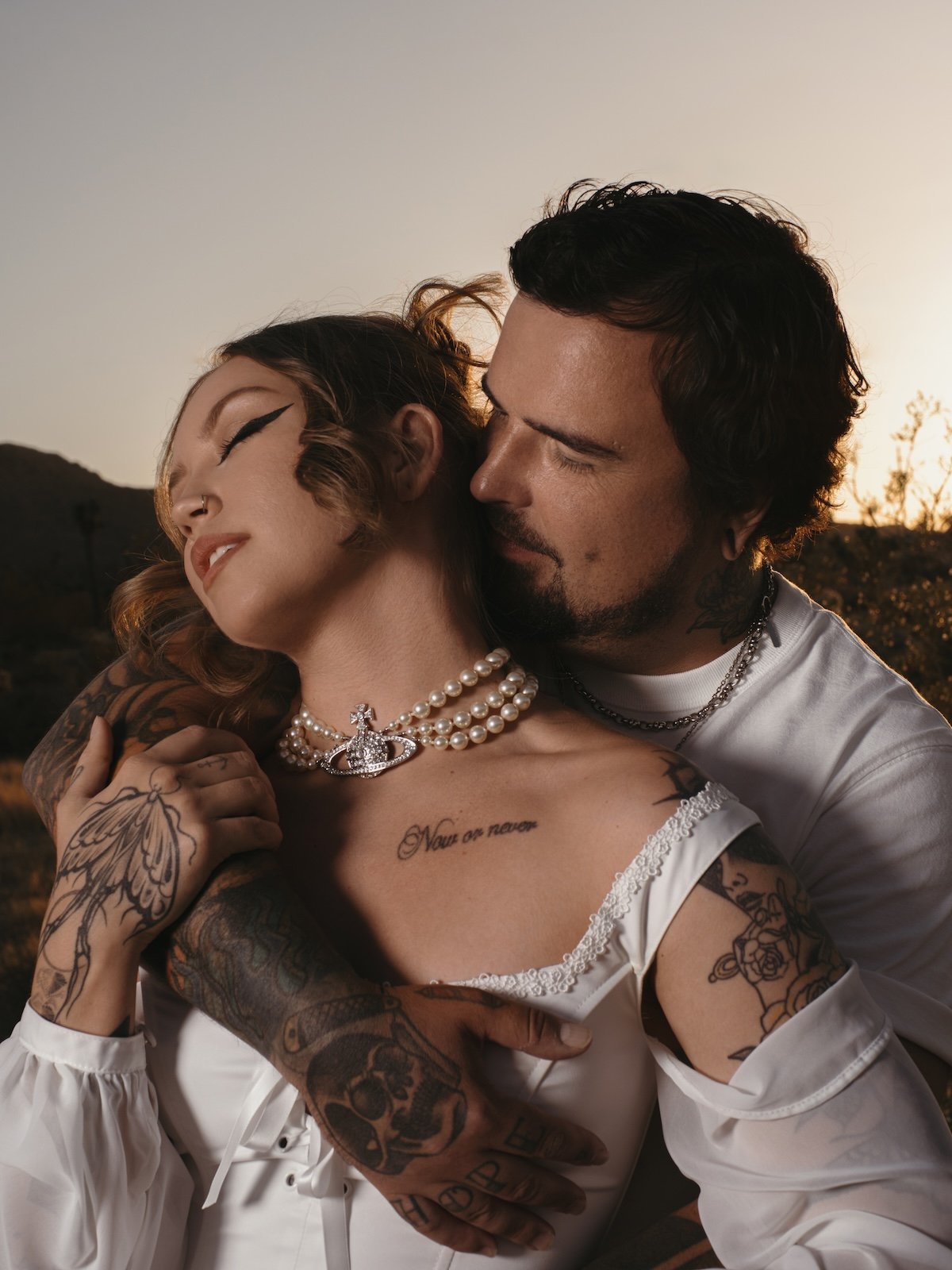 A groom and bride embrace outdoors during sunset, with the woman wearing pearl jewelry and tattoos, and the man close to her face.