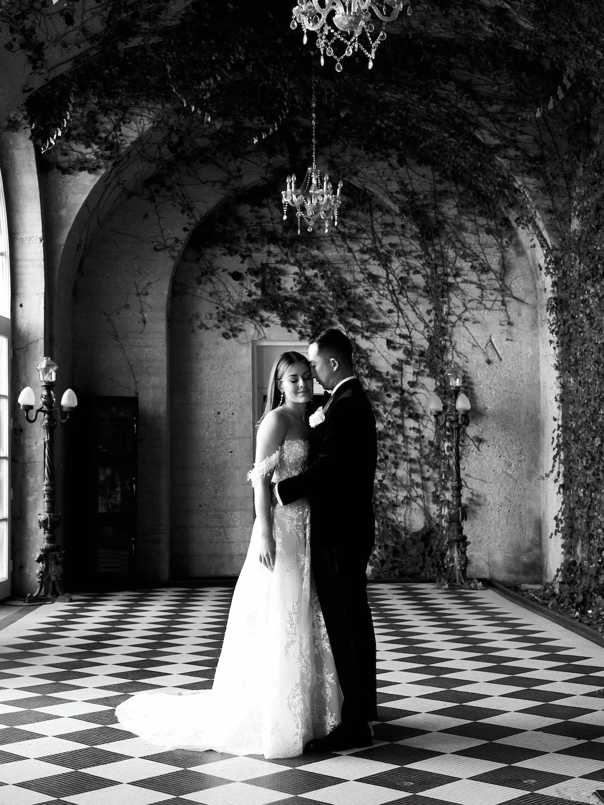 A bride and groom share a dance in a beautifully decorated indoor space with chandeliers, tall lamps, and ivy-covered walls, captured in black and white.