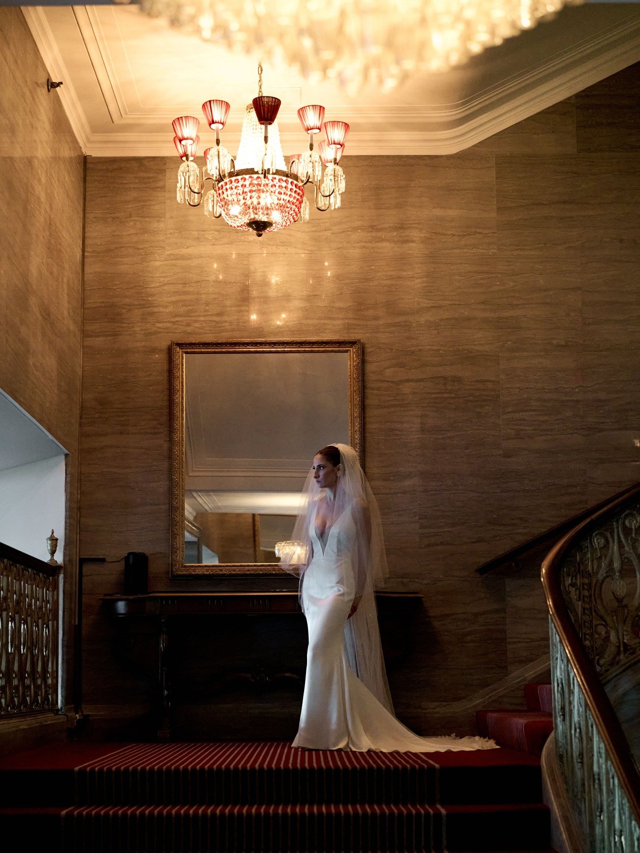 A bride in a white wedding dress with a veil standing on a staircase in an elegant, warmly lit interior with a chandelier above and a framed mirror behind her.