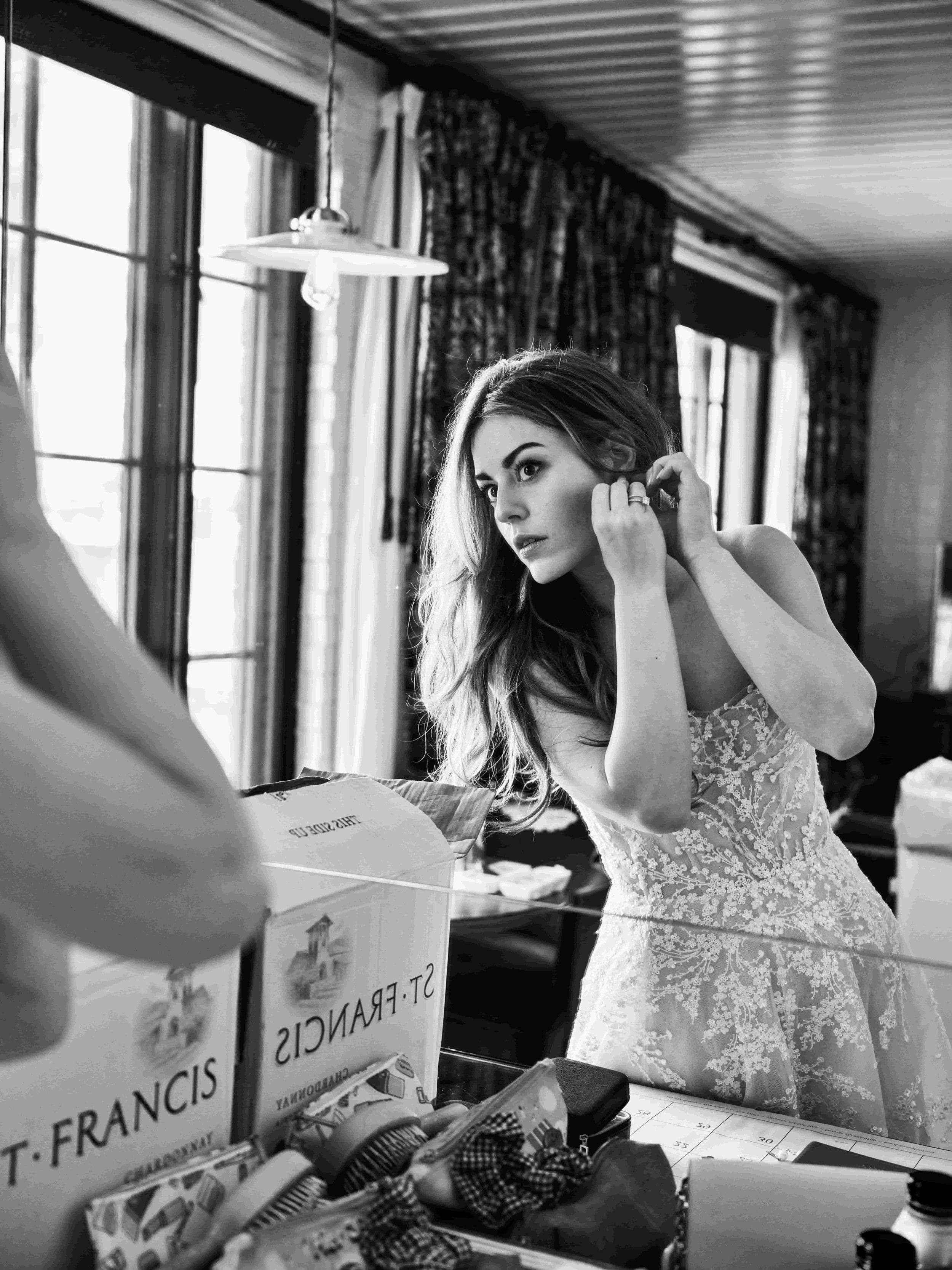 A woman wearing a wedding dress is looking into a mirror and adjusting her earring in a room with large windows and curtains.