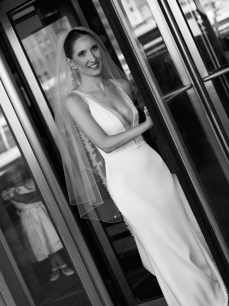Black and white photo of a smiling bride in a wedding dress with a veil, standing inside a glass elevator or elevator door.