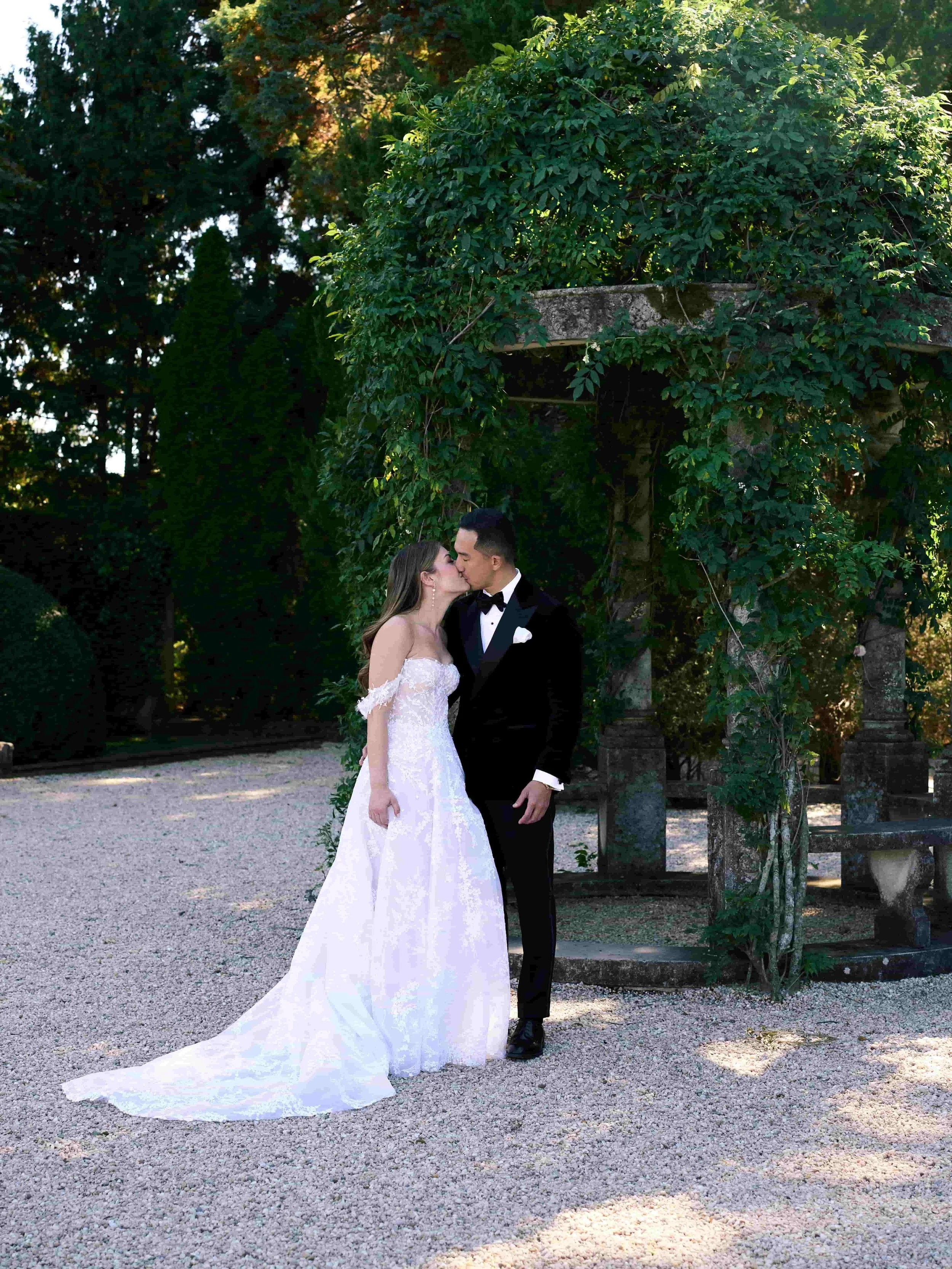 A bride in a white wedding gown and a groom in a black tuxedo share a kiss outdoors under an arbor covered with green vines.