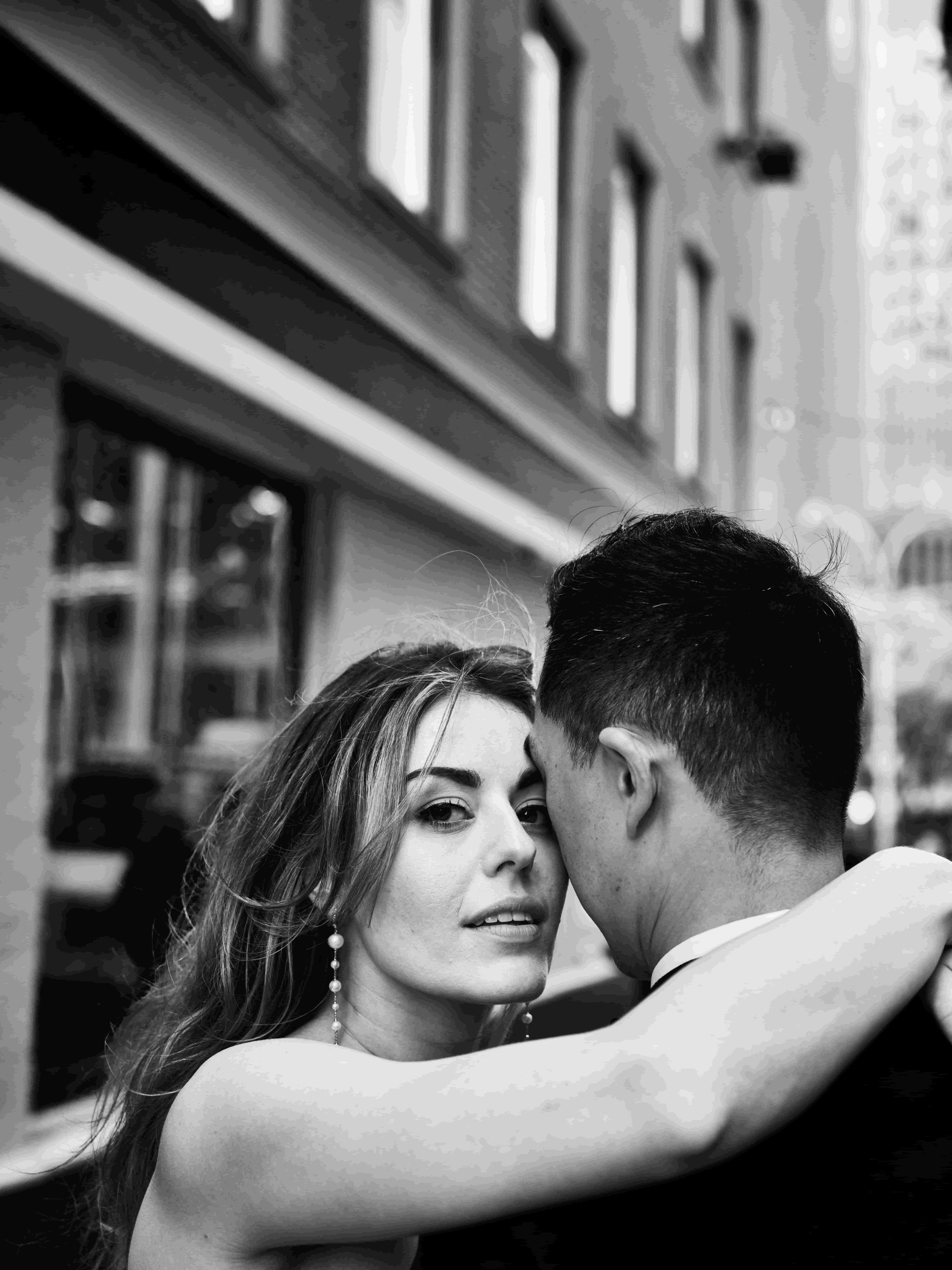 A black and white photo of a woman and man dancing closely, with the woman looking at the camera and the man with his back to the camera, in an outdoor setting with buildings in the background.