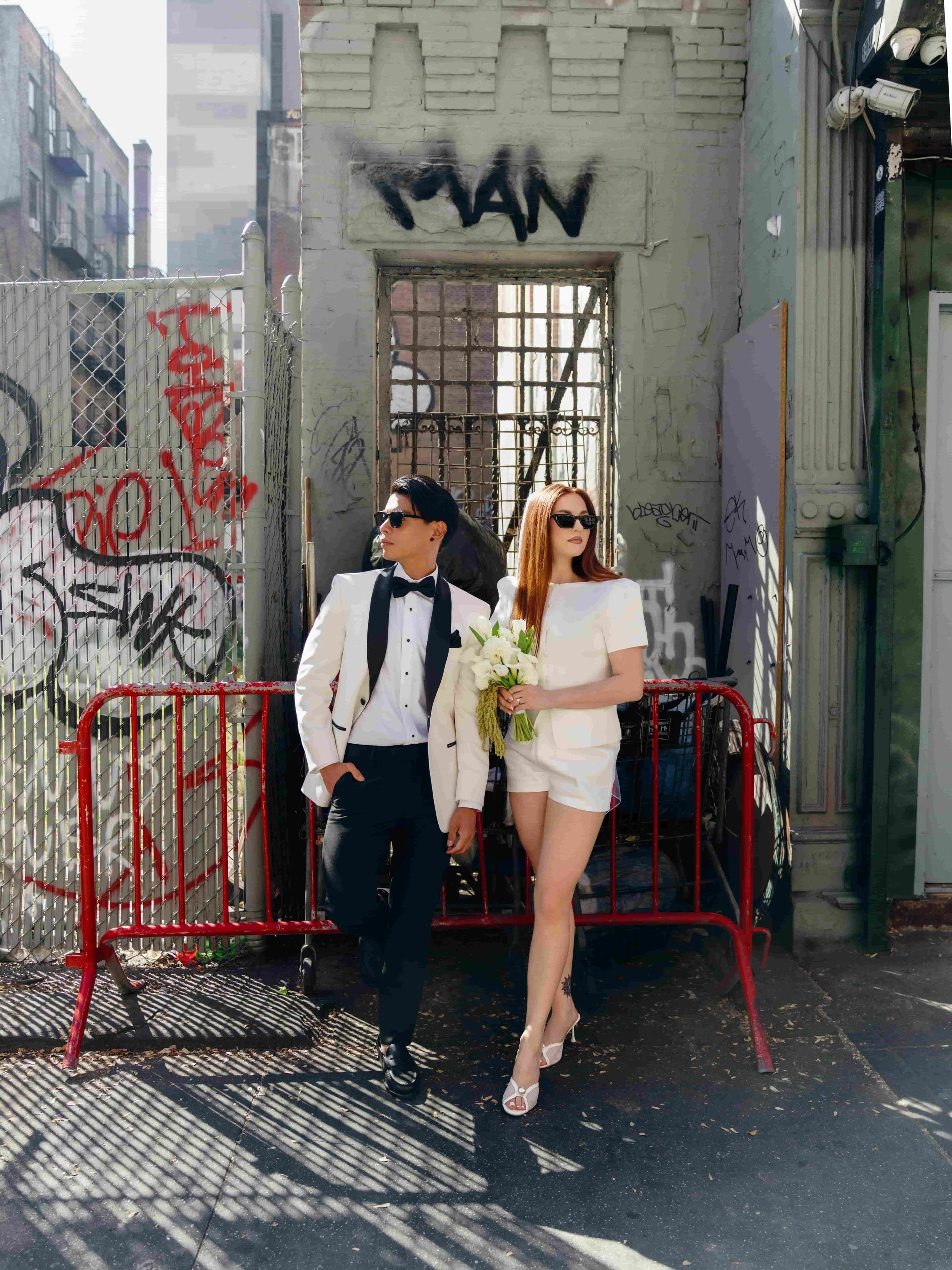 A stylish couple dressed in formal white outfits pose outdoors against a graffiti-covered wall, with the woman holding a bouquet of white flowers.