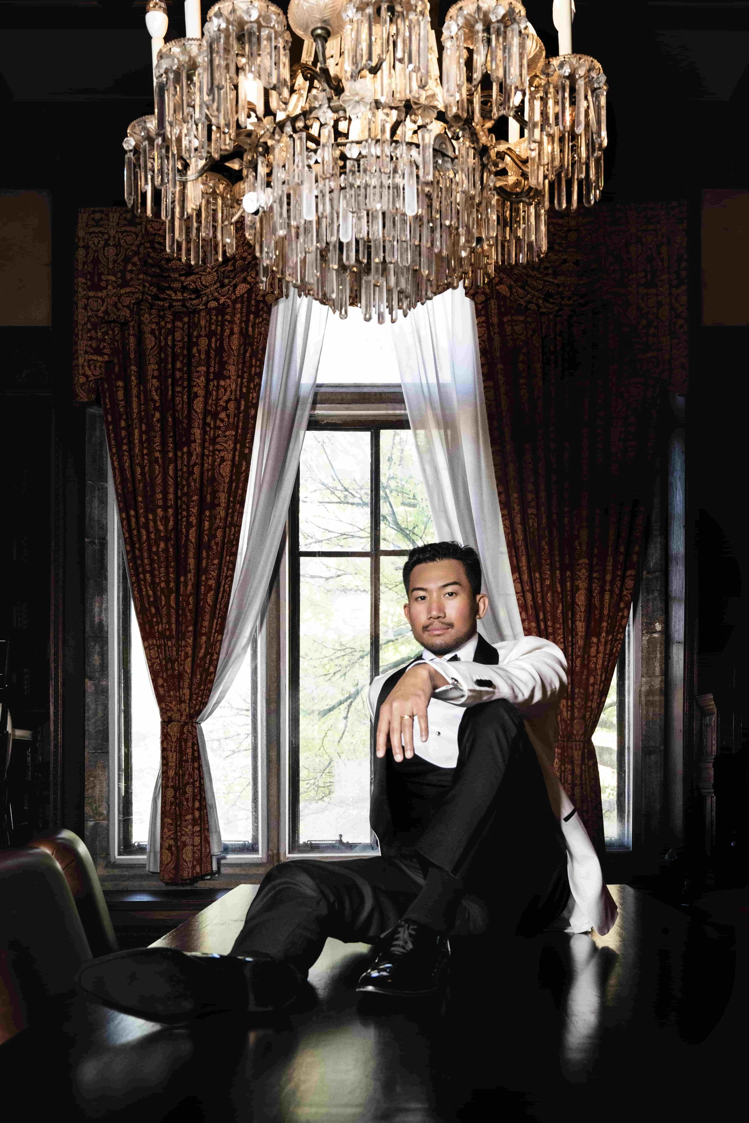 A groom in a tuxedo sitting on a dark wooden table in a luxurious room with a large chandelier, tall windows with curtains, and outdoor view of trees.