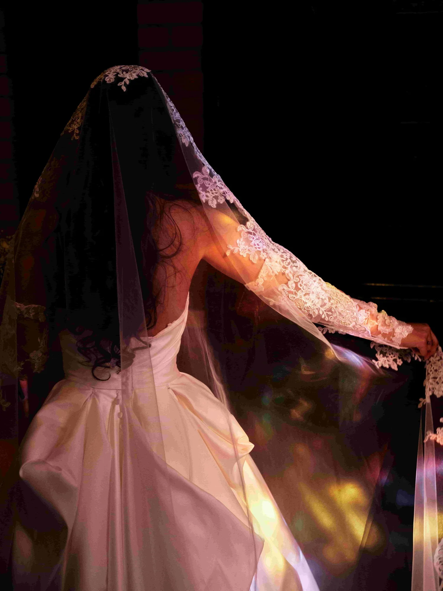 A woman in a white wedding dress with long lace sleeves, wearing a black veil, is adjusting her dress in a dimly lit room.