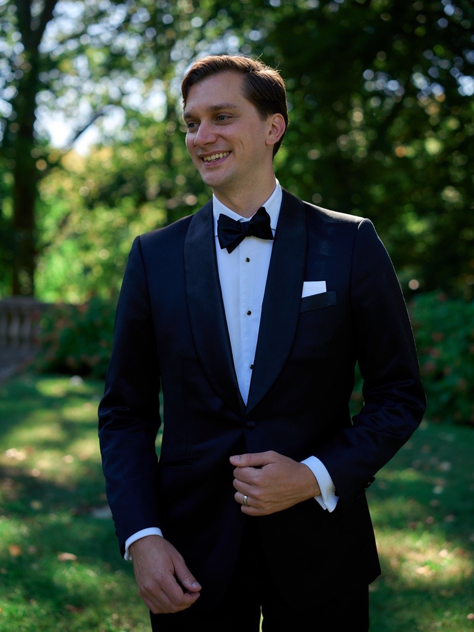 A man in a black tuxedo with a bowtie, standing outdoors with green trees in the background, smiling.