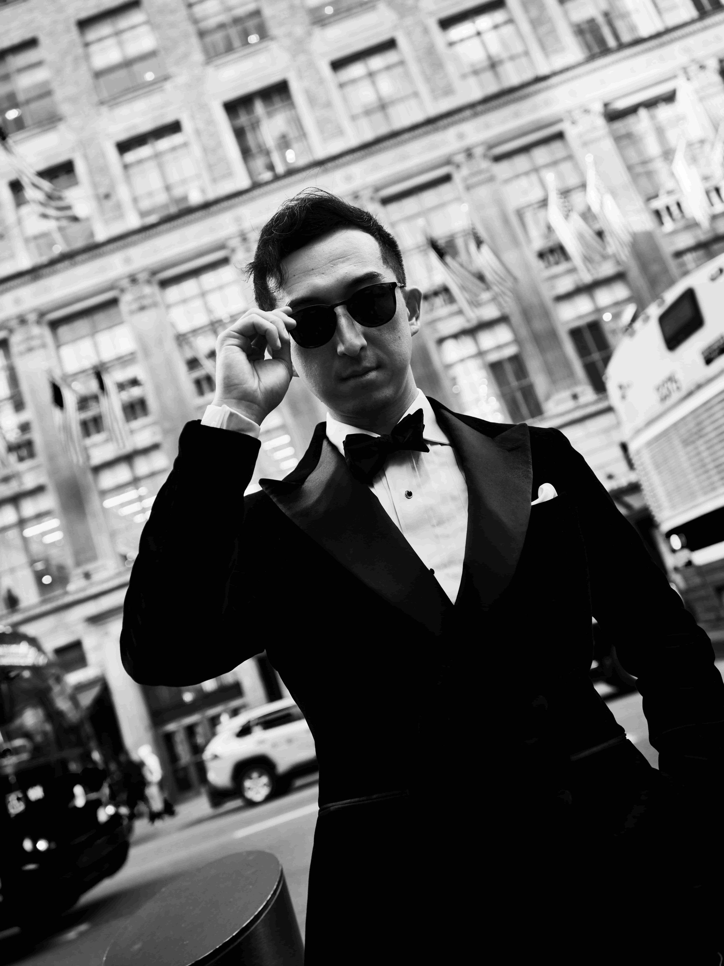 A man dressed in a tuxedo and sunglasses standing on a city street with buildings and cars in the background.