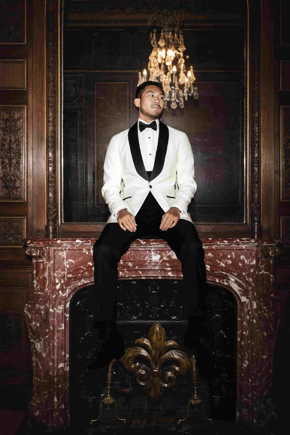 A man dressed in a white tuxedo jacket with black lapels, black pants, and a black bow tie, sitting on a marble fireplace mantel in an elegant, ornate room.
