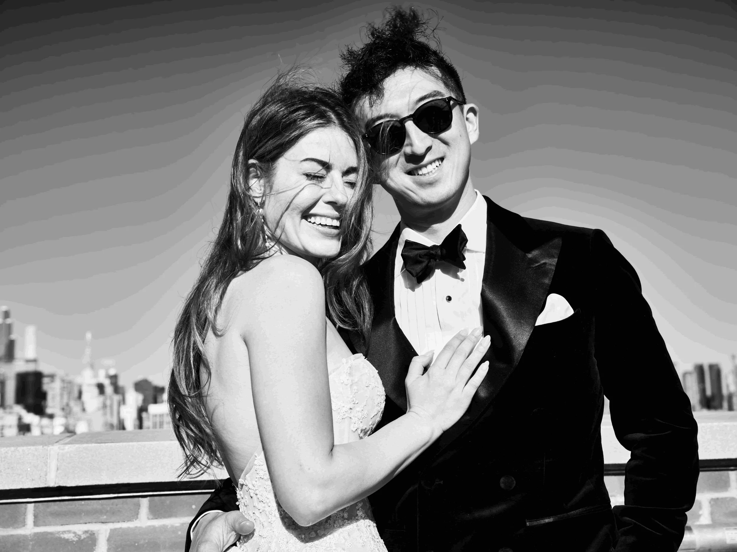 Black and white photo of a smiling couple on a rooftop, the woman in a strapless dress and the man in a suit with sunglasses.