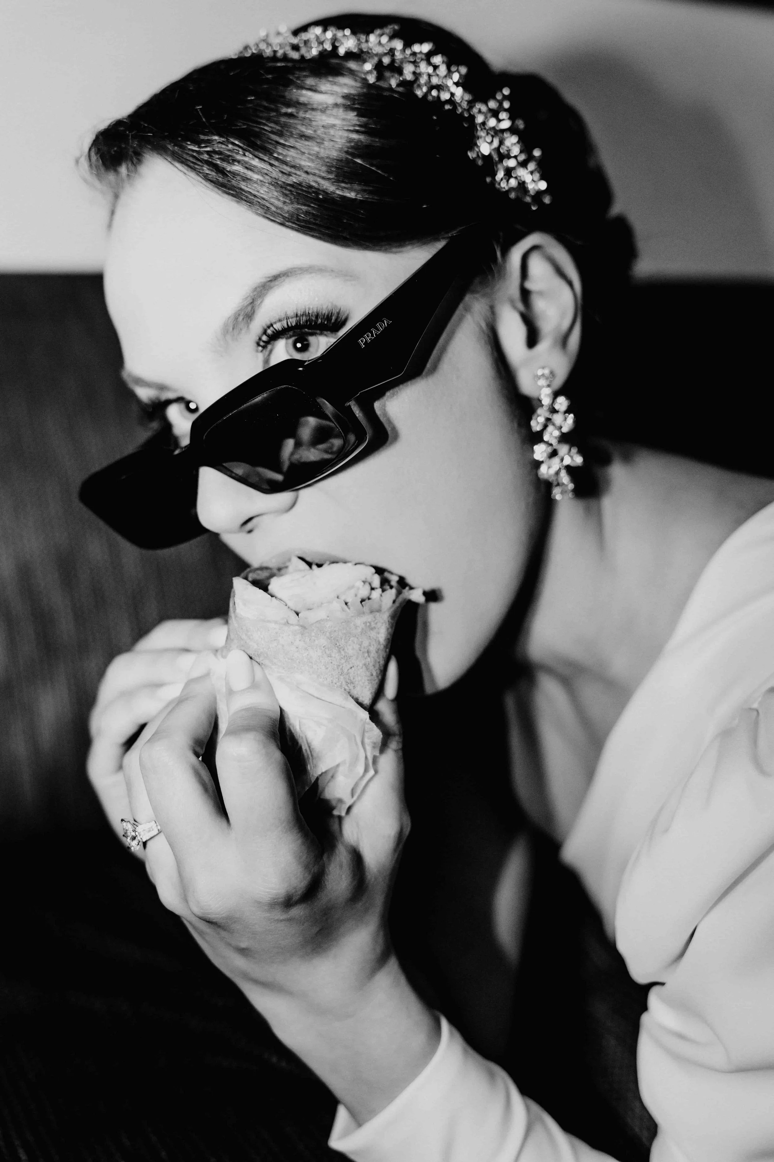 A woman wearing large Prada sunglasses, diamond earrings, and a ring on her left hand eating a wrap or burrito, with her hair styled neatly and adorned with a sparkly hair accessory.
