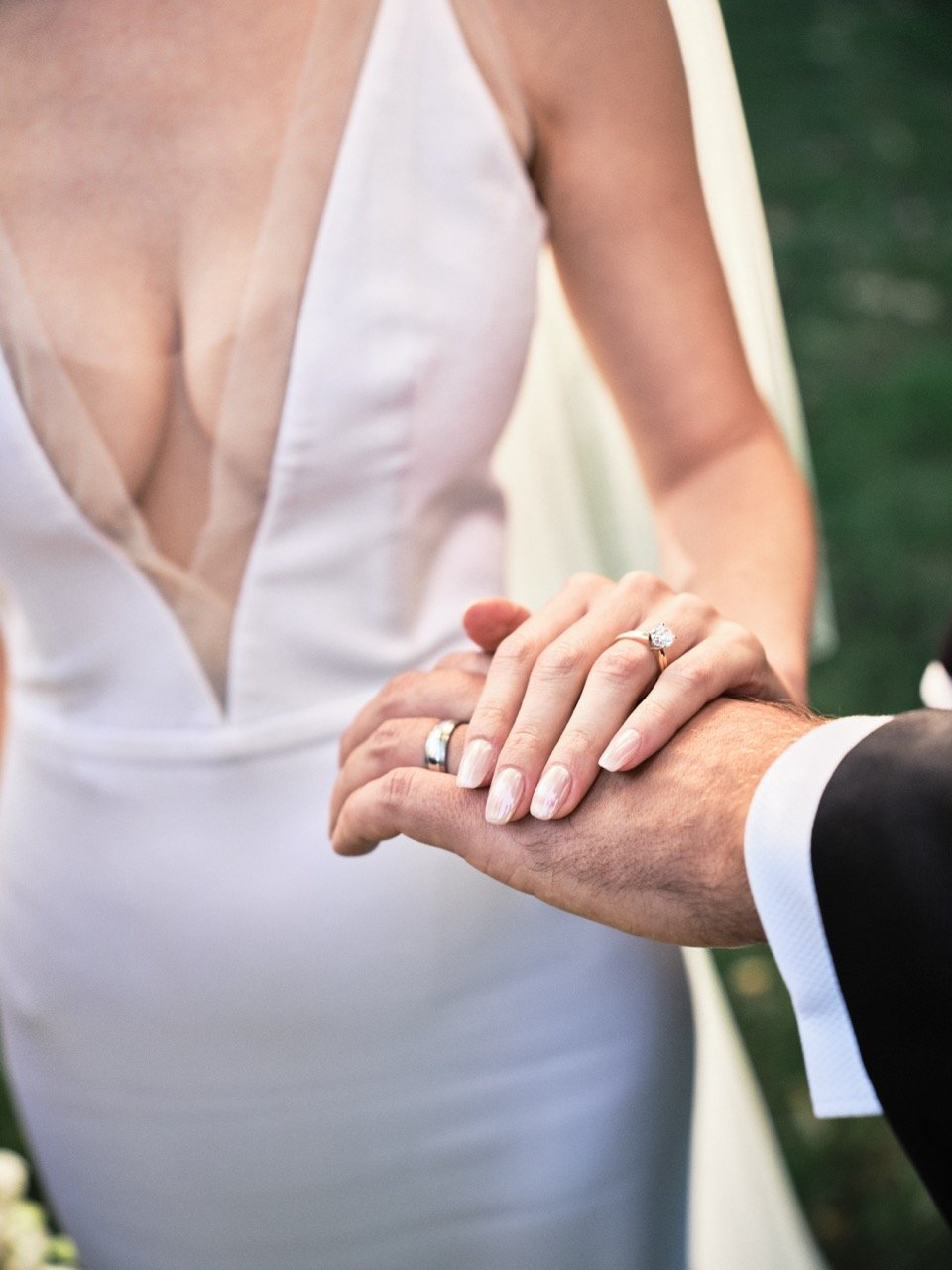 Close-up of a bride and groom holding hands during a wedding ceremony, with her wearing a wedding ring and a wedding dress, and him in a tuxedo.