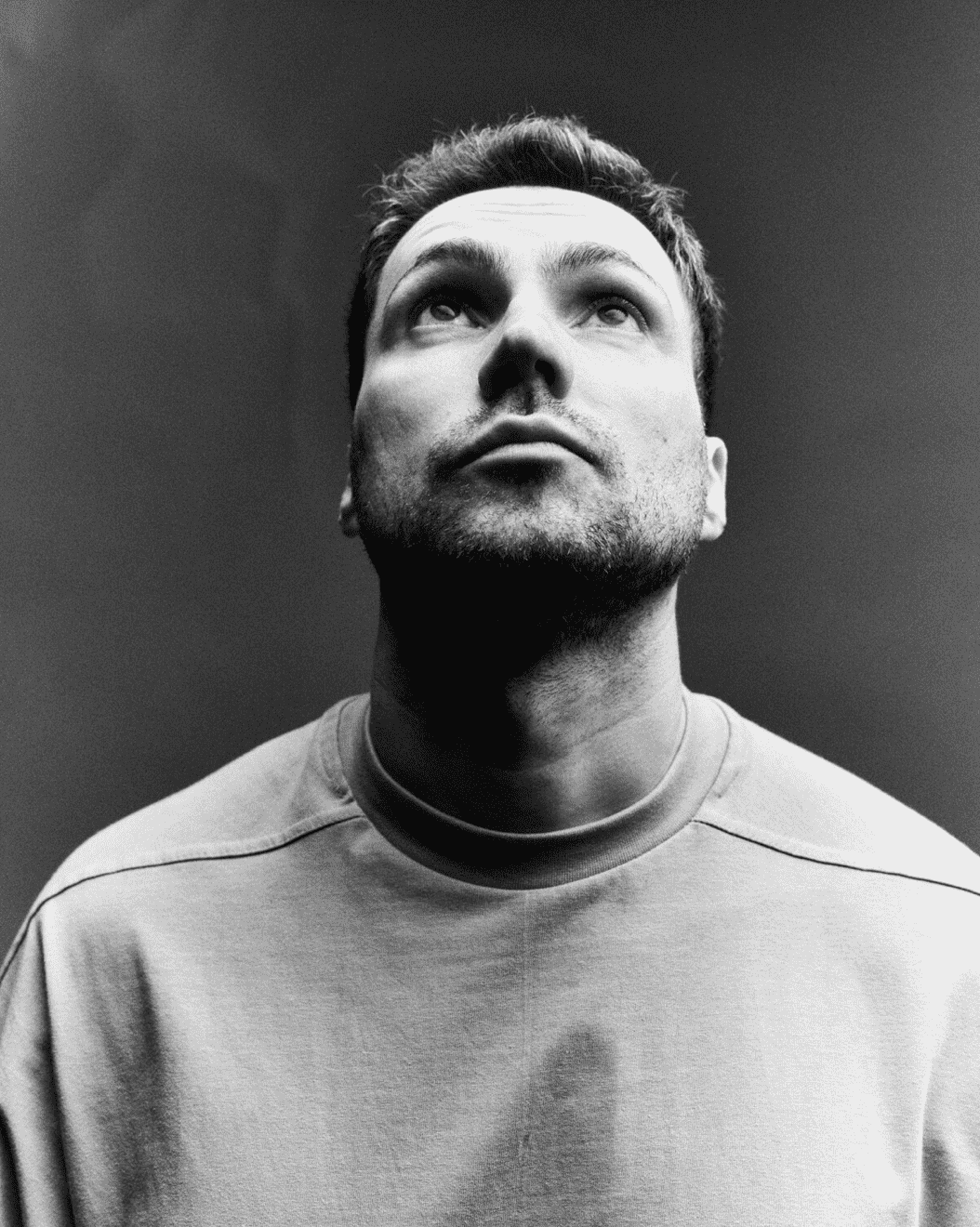 A black and white portrait of a man looking upward, wearing a light-colored sweatshirt, set against a dark background.