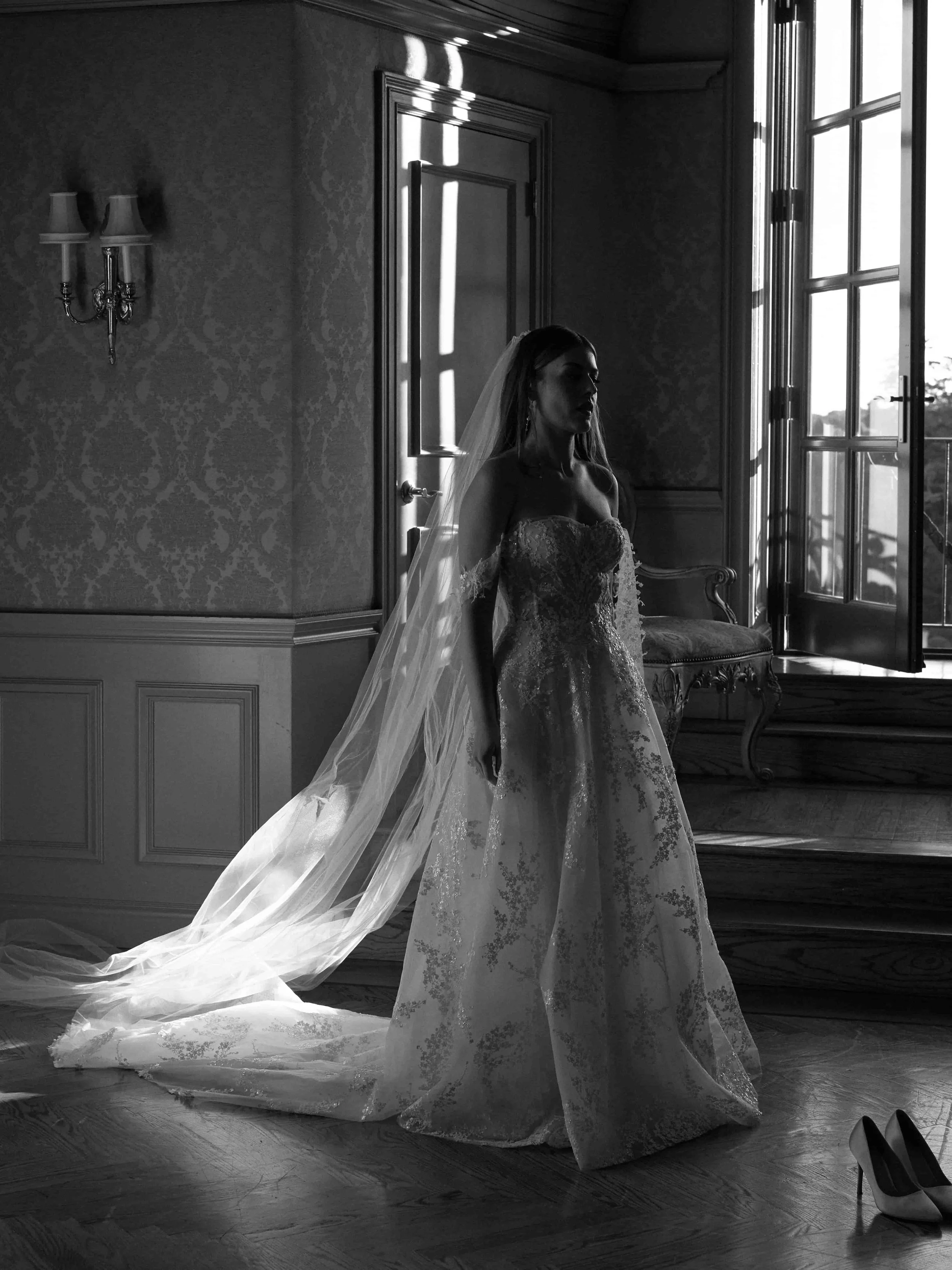 A bride in a wedding dress with a long train stands inside a room near an open window, with sunlight coming through and casting shadows on the wall. A pair of high heels is on the floor nearby.