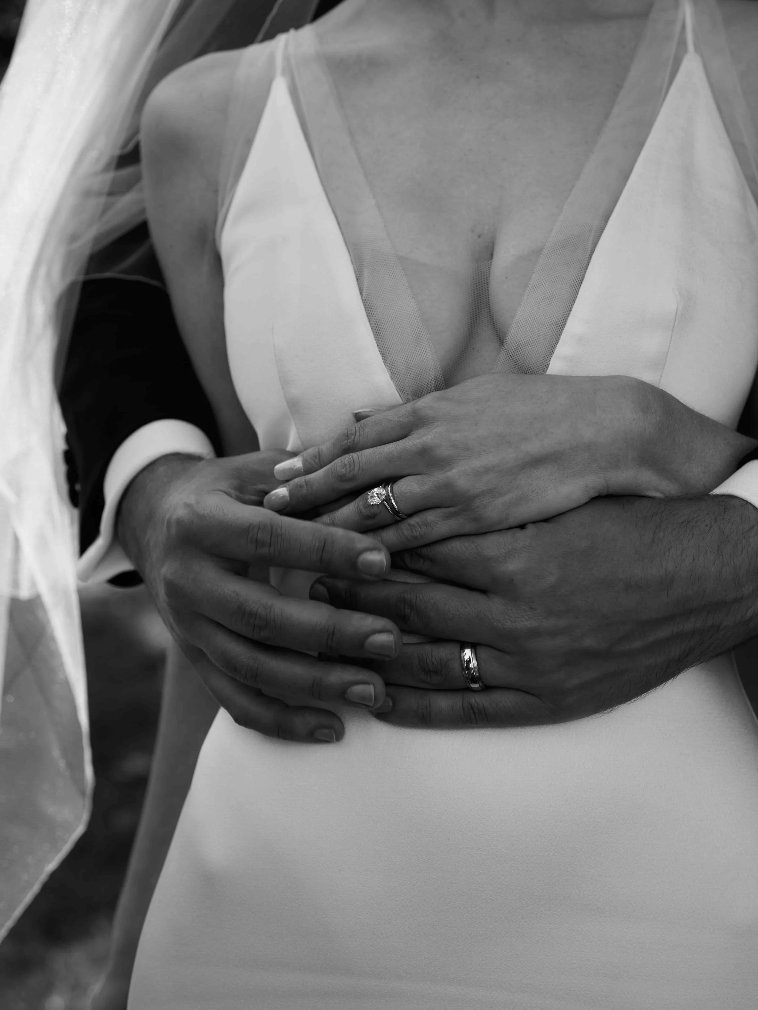 Close-up of a couple embracing, wearing wedding rings. The bride is in a white dress and veil, and the groom's hand is around her waist.