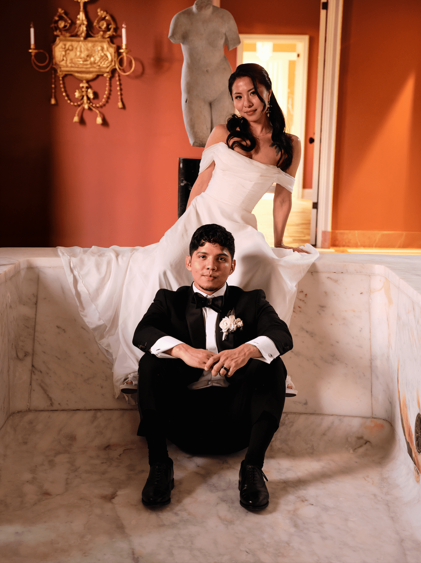 A bride in a white wedding dress sitting on a marble ledge, with her hand resting on her lap, and a groom in a black tuxedo sitting on the ground in front of her, both posing indoors with classical decor and a statue in the background.