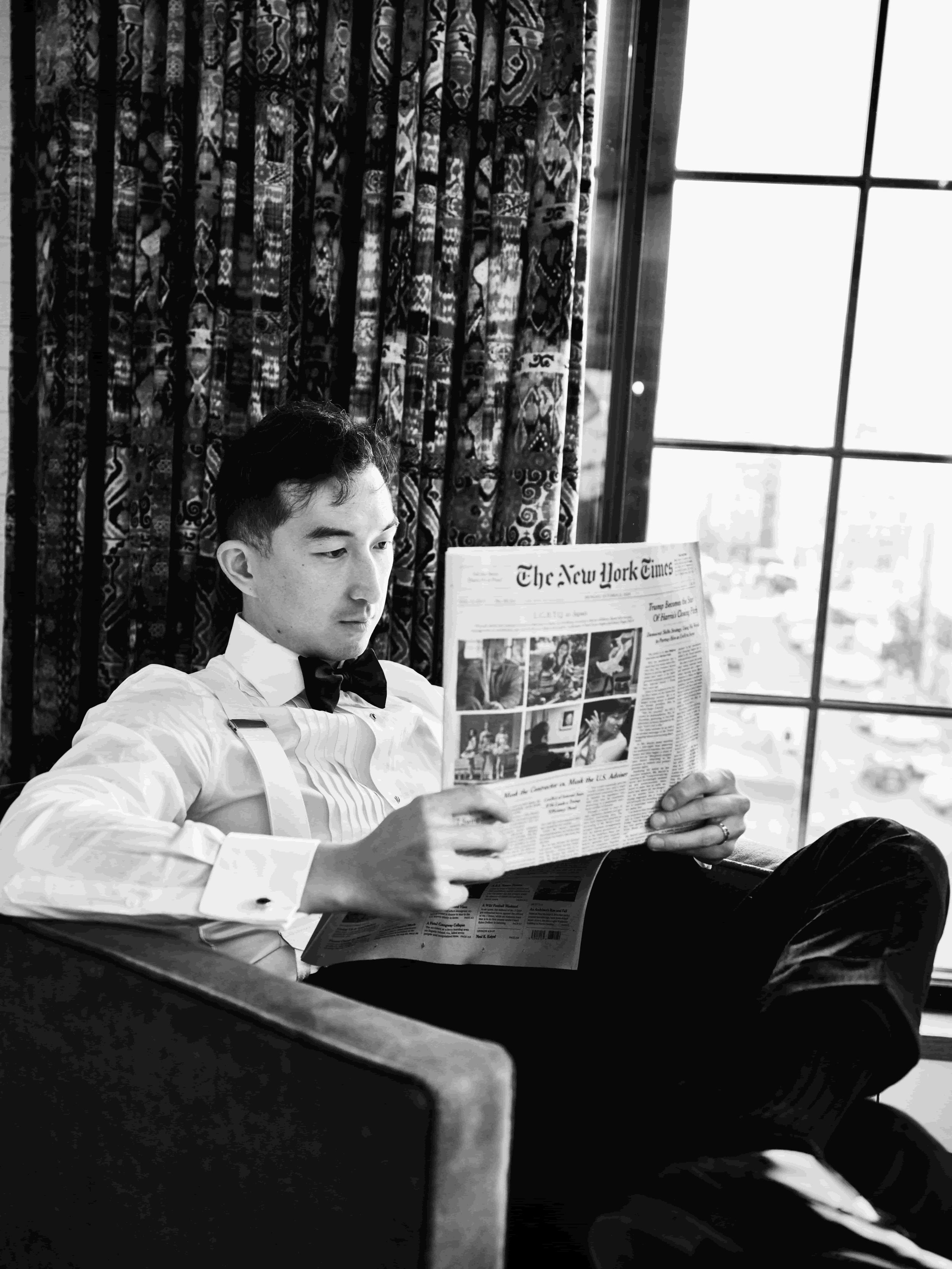 A young man dressed in a white shirt, black bow tie, and suspenders, sitting in a chair reading a copy of The New York Times, with a window and patterned curtains in the background.