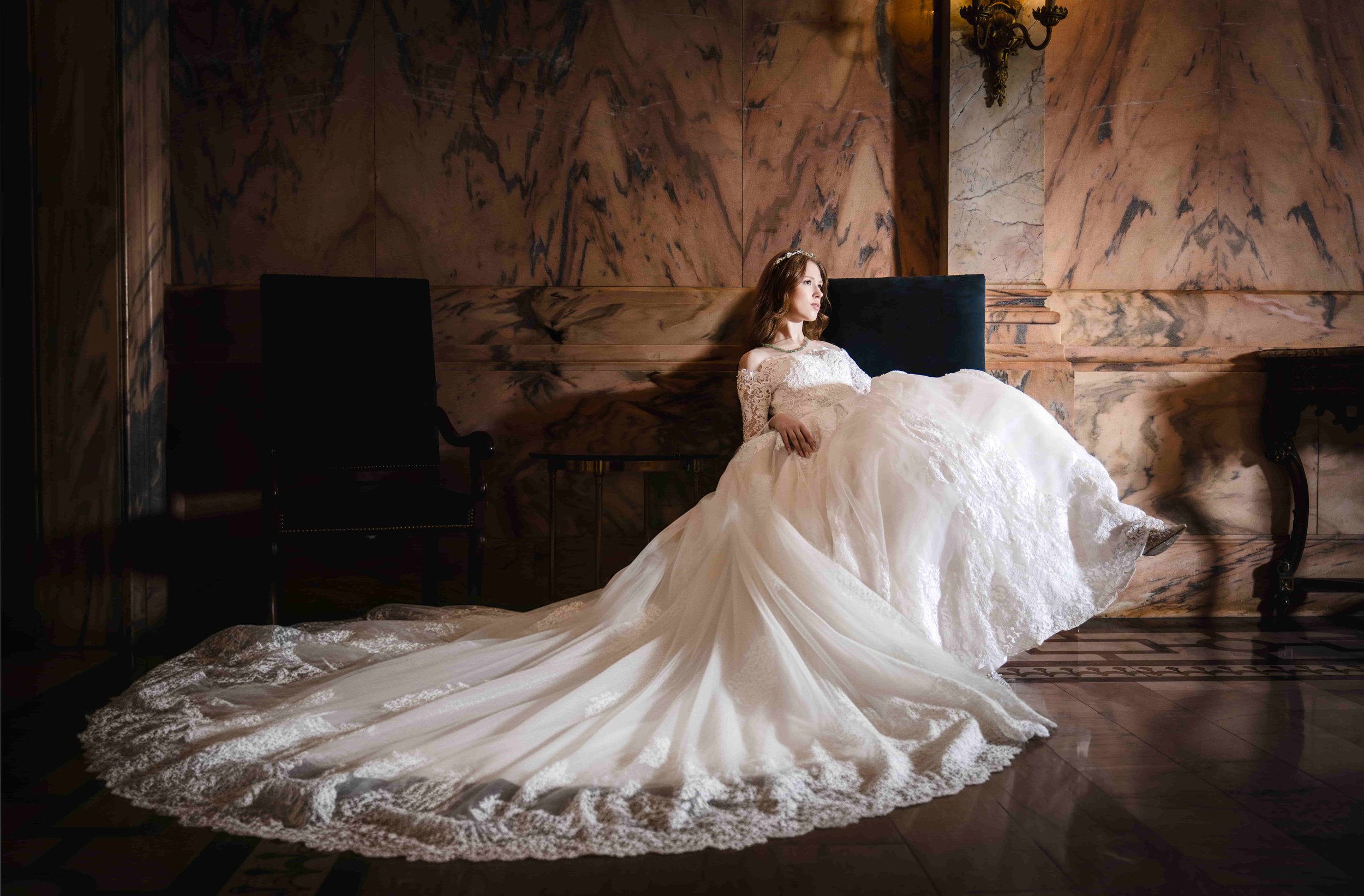 A bride in a white wedding gown with lace sleeves sitting on a bench in an elegant, dimly lit interior with marble walls and wooden flooring.
