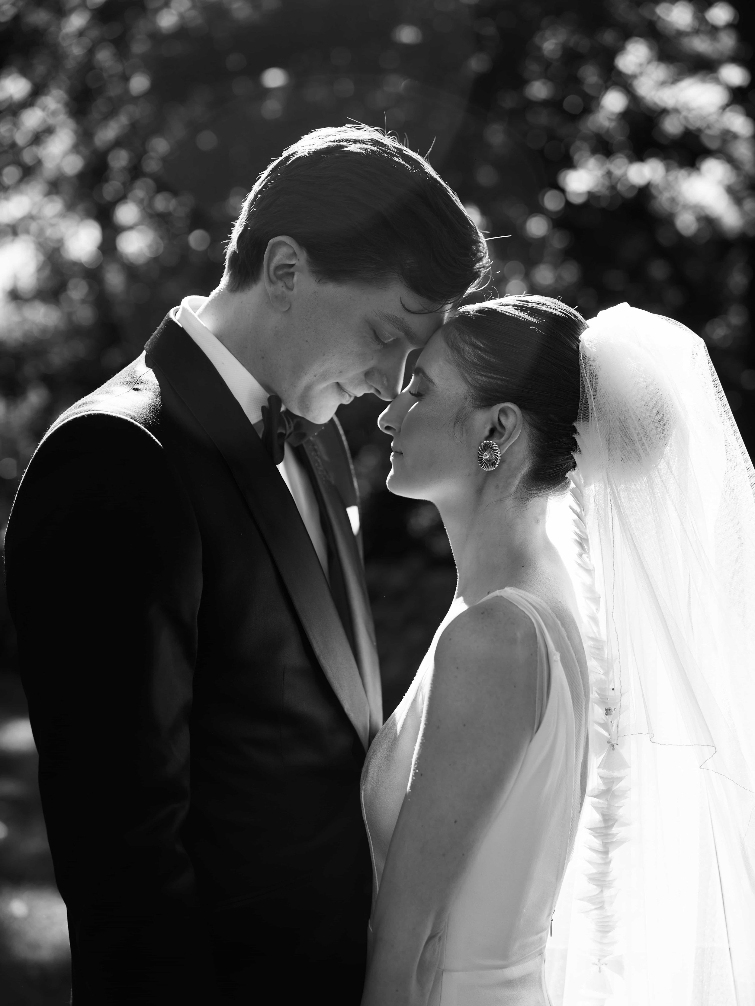 Black and white photo of a bride and groom in an intimate moment, with their foreheads touching and eyes closed, outdoors with trees in the background.