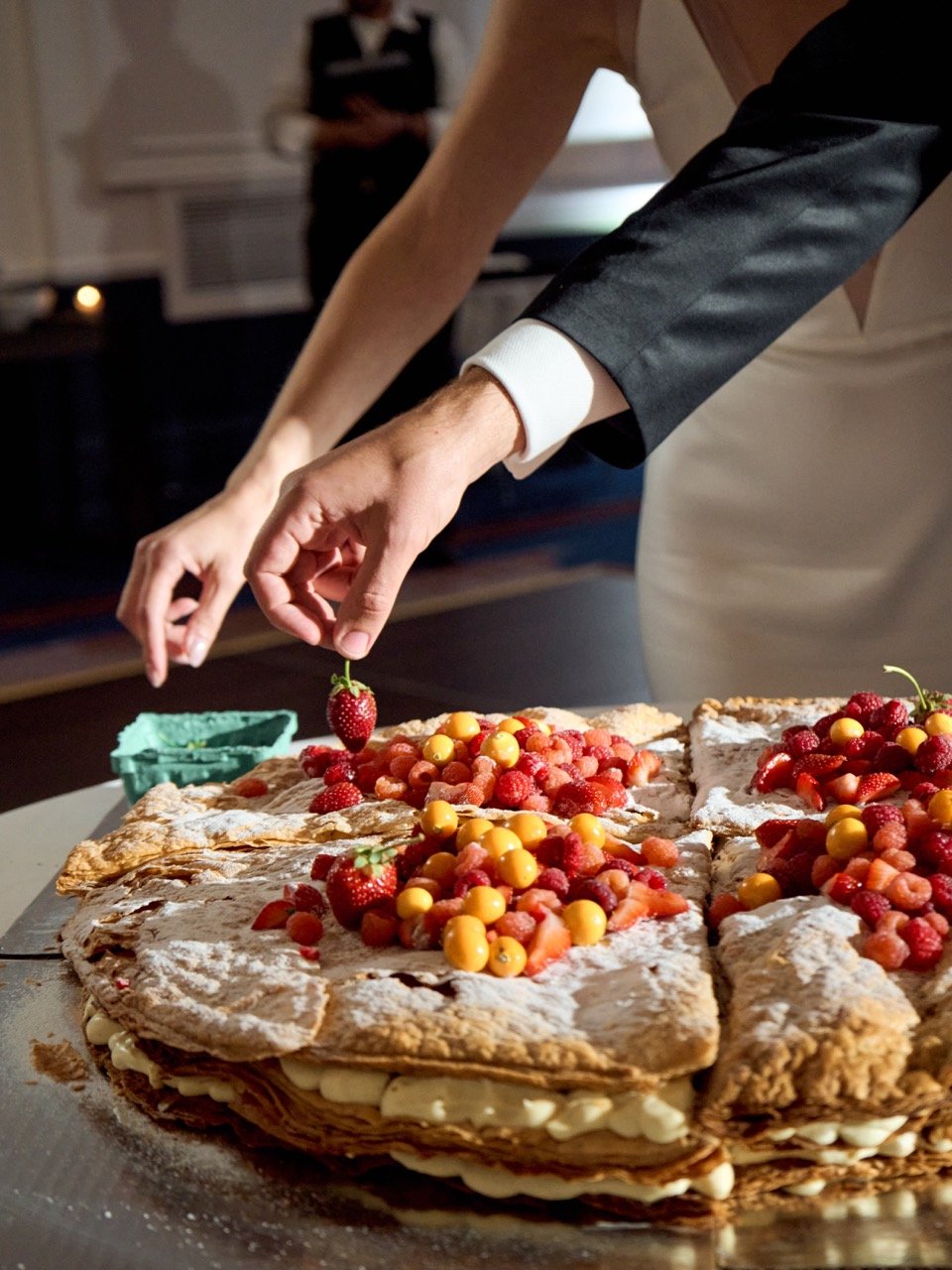 A person in formal attire is slicing a large layered cake topped with strawberries, raspberries, and yellow berries at a celebration or event.