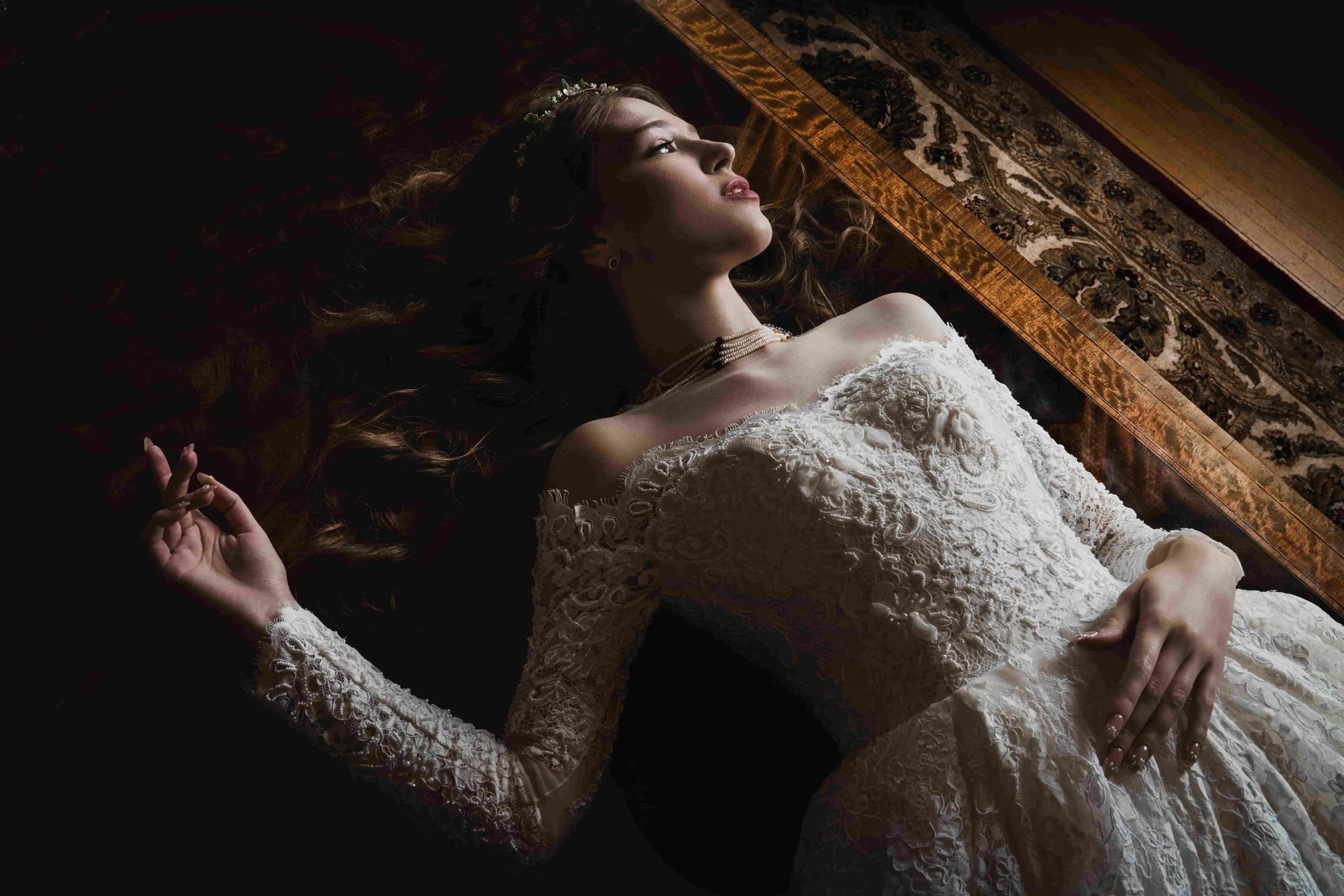 A woman lying on the floor in a white lace dress, with her hair spread out, looking up, on a dark surface near a patterned rug.
