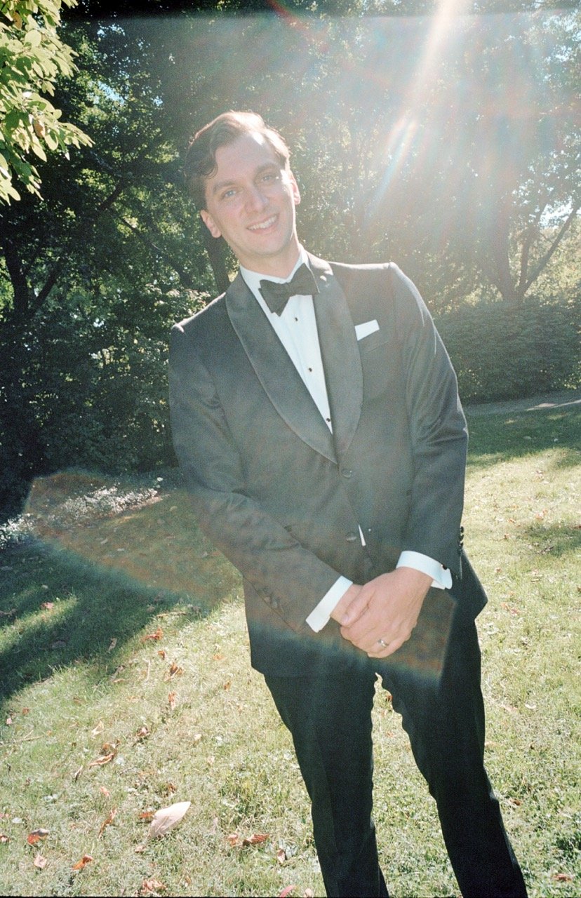 A man in a tuxedo standing outdoors with trees in the background, sunlight shining on him, smiling and posing for the photo.
