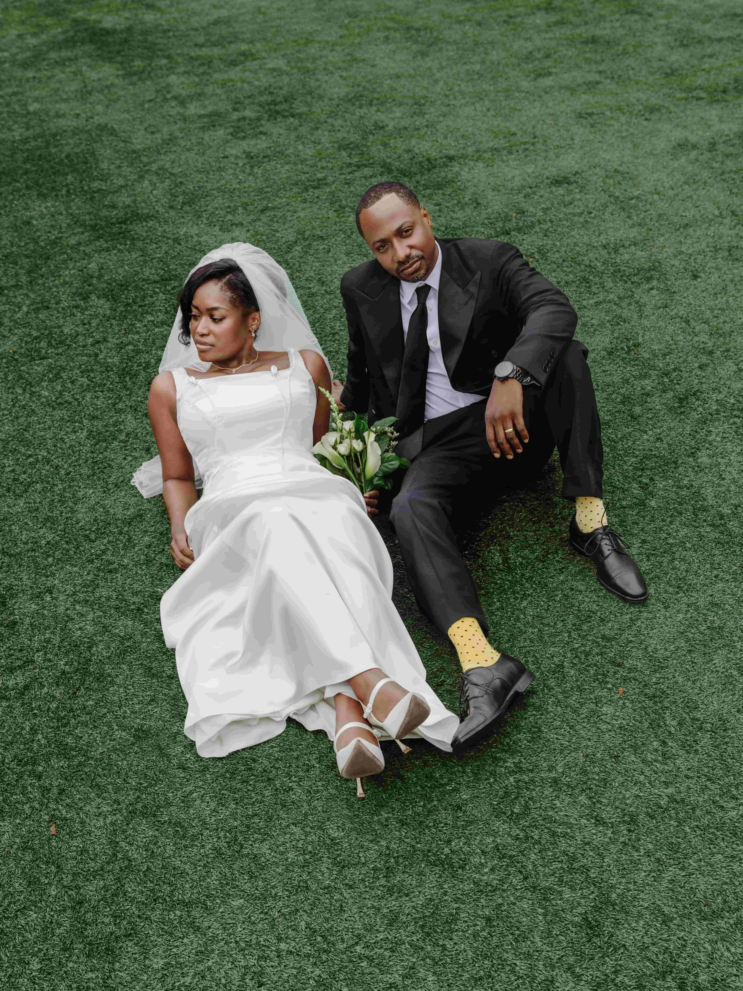 A bride in a white wedding dress and a groom in a black suit sitting on green grass, with the bride holding a bouquet of white flowers and the groom wearing yellow polka dot socks.