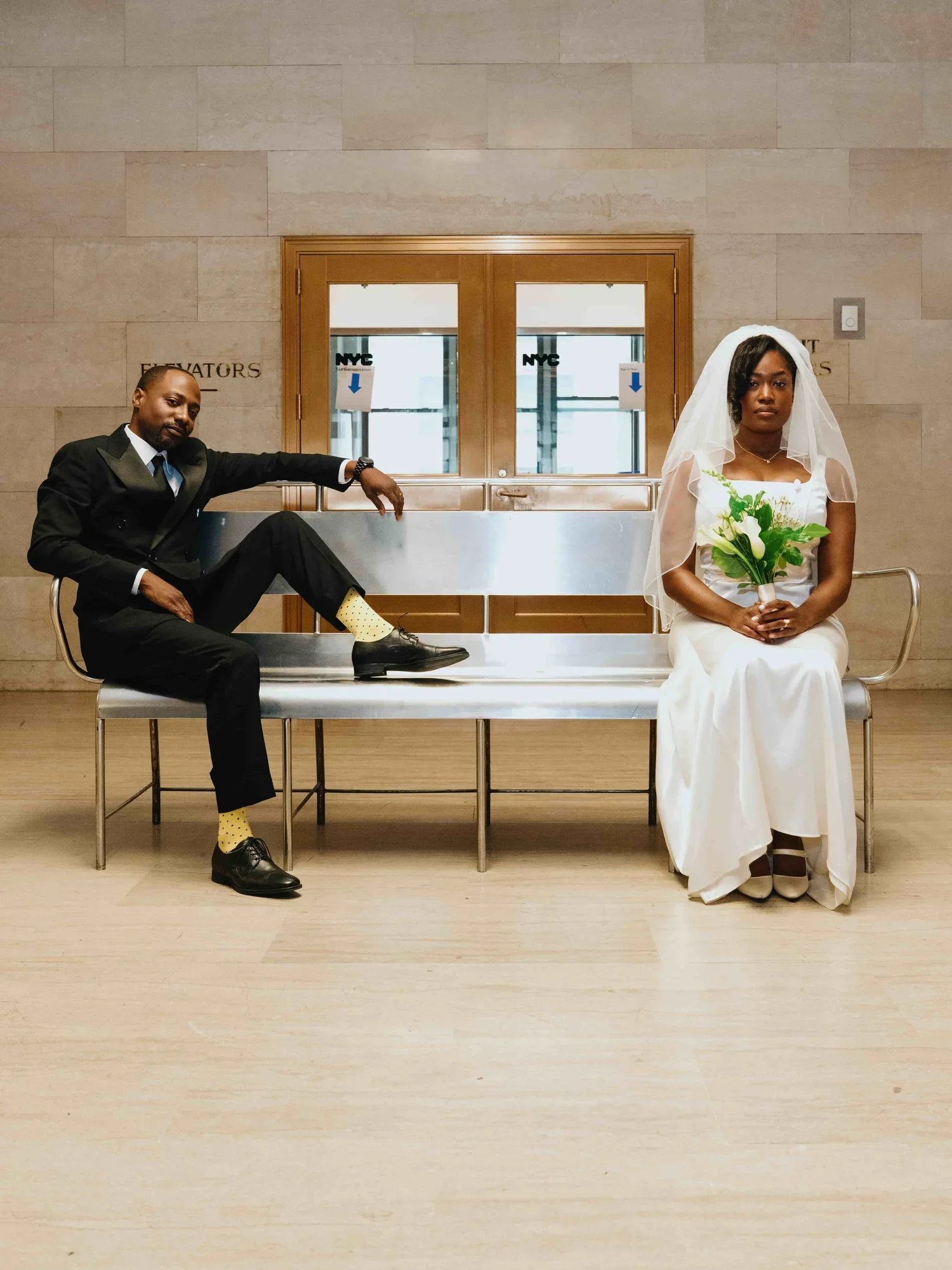 A man in a black suit with yellow socks and black shoes sitting on a bench next to a woman in a white wedding dress with a veil, holding a bouquet of white flowers, inside a building with a beige wall and wooden doors.