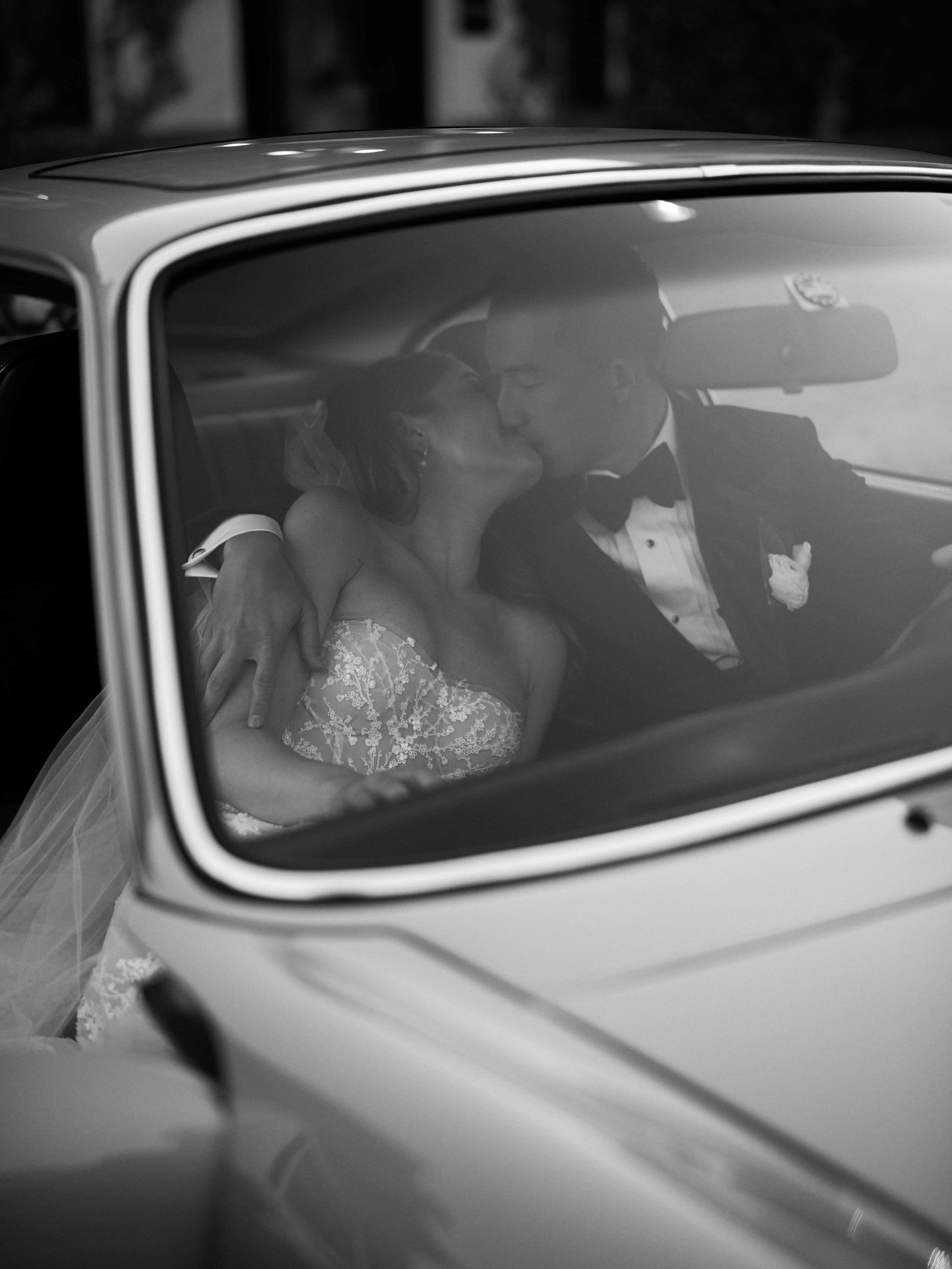 A black and white photo of a bride and groom sharing a kiss inside a car on their wedding day