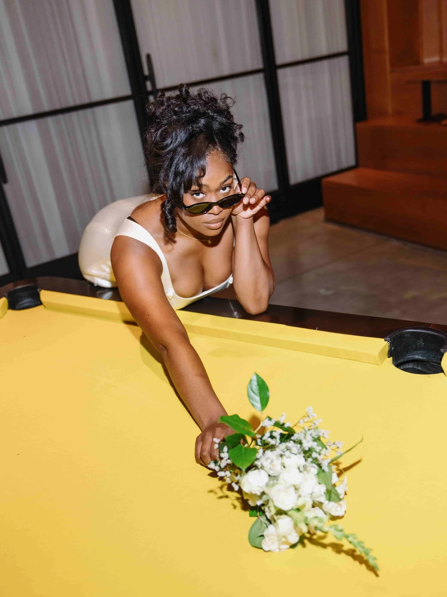 A woman with curly hair and medium skin tone leaning on a yellow ping pong table, wearing sunglasses, a white dress, and holding a bouquet of white flowers, looking at the camera.