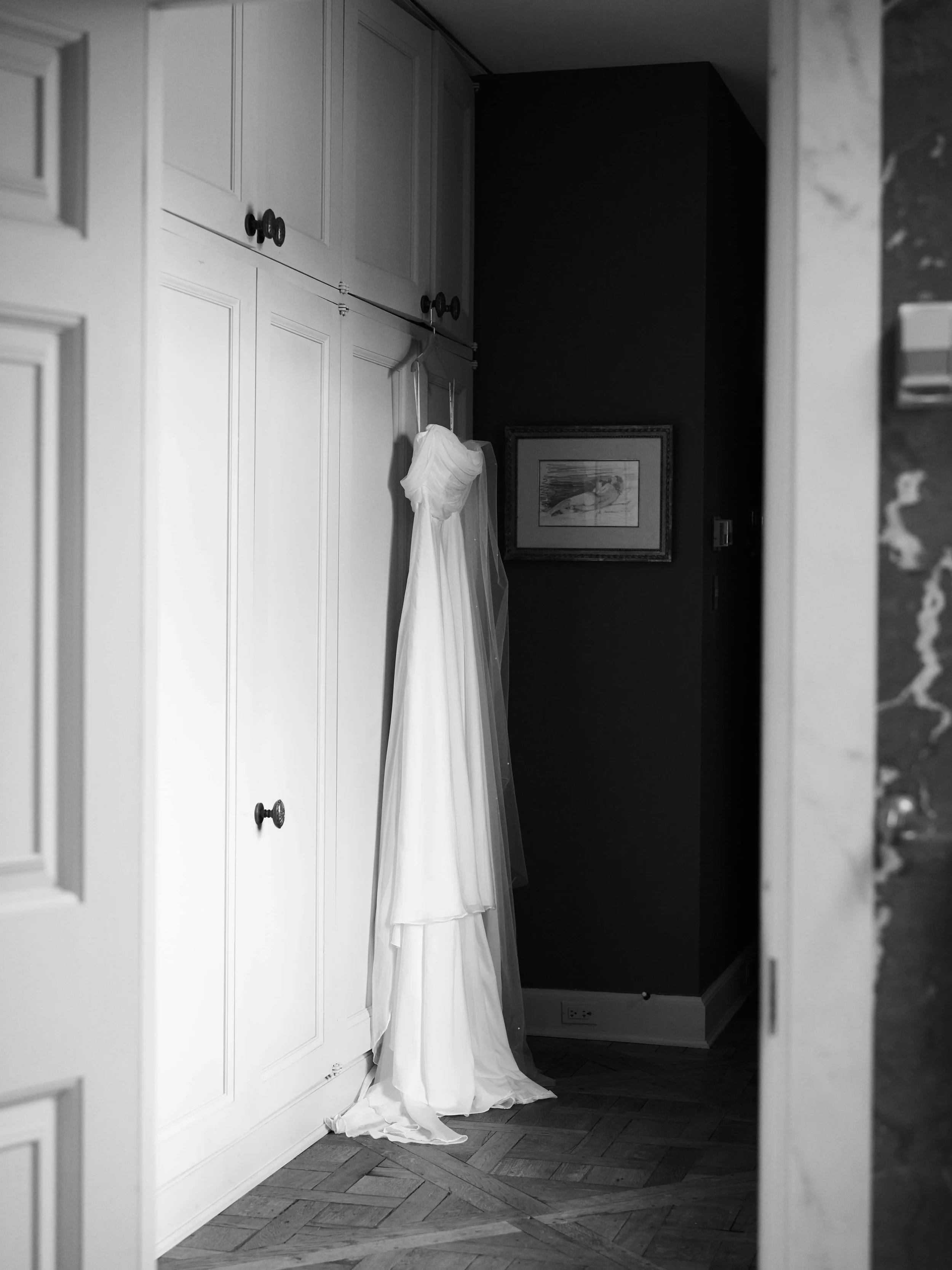 A wedding dress hanging on a door in a room with white cabinetry, dark walls, framed art, wooden floor, and a marble countertop visible on the right.