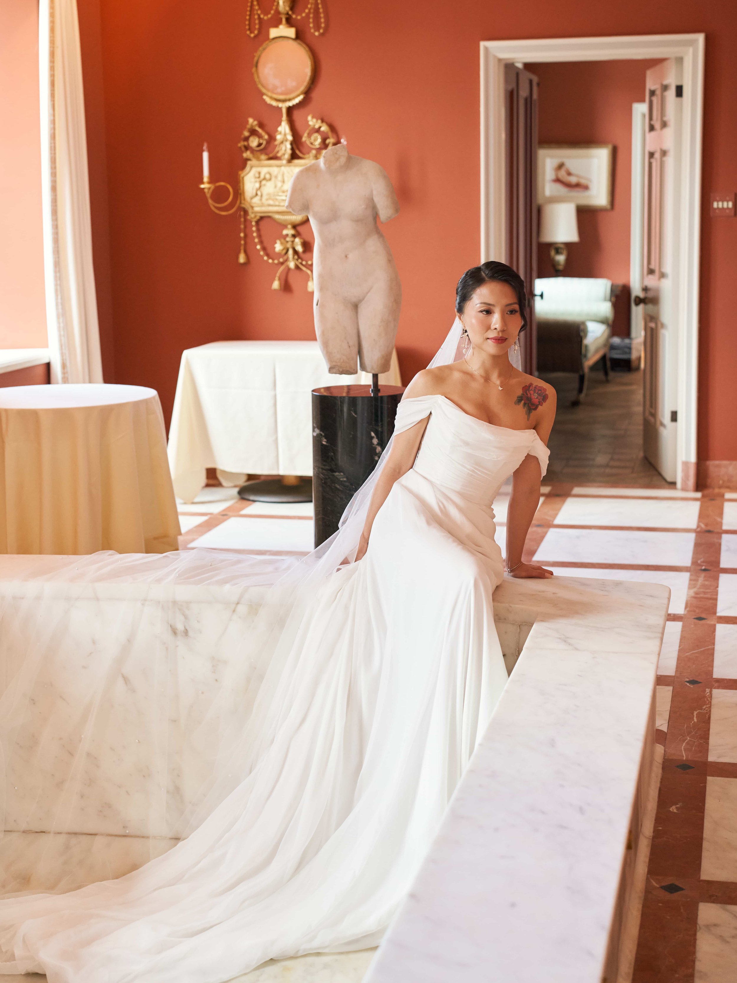 A bride in a white wedding dress with off-shoulder sleeves, sitting on the edge of a marble bathtub in an elegant room with red walls. There is a classical sculpture of a male torso on a stand, an ornate wall sconce, and a doorway leading to another 