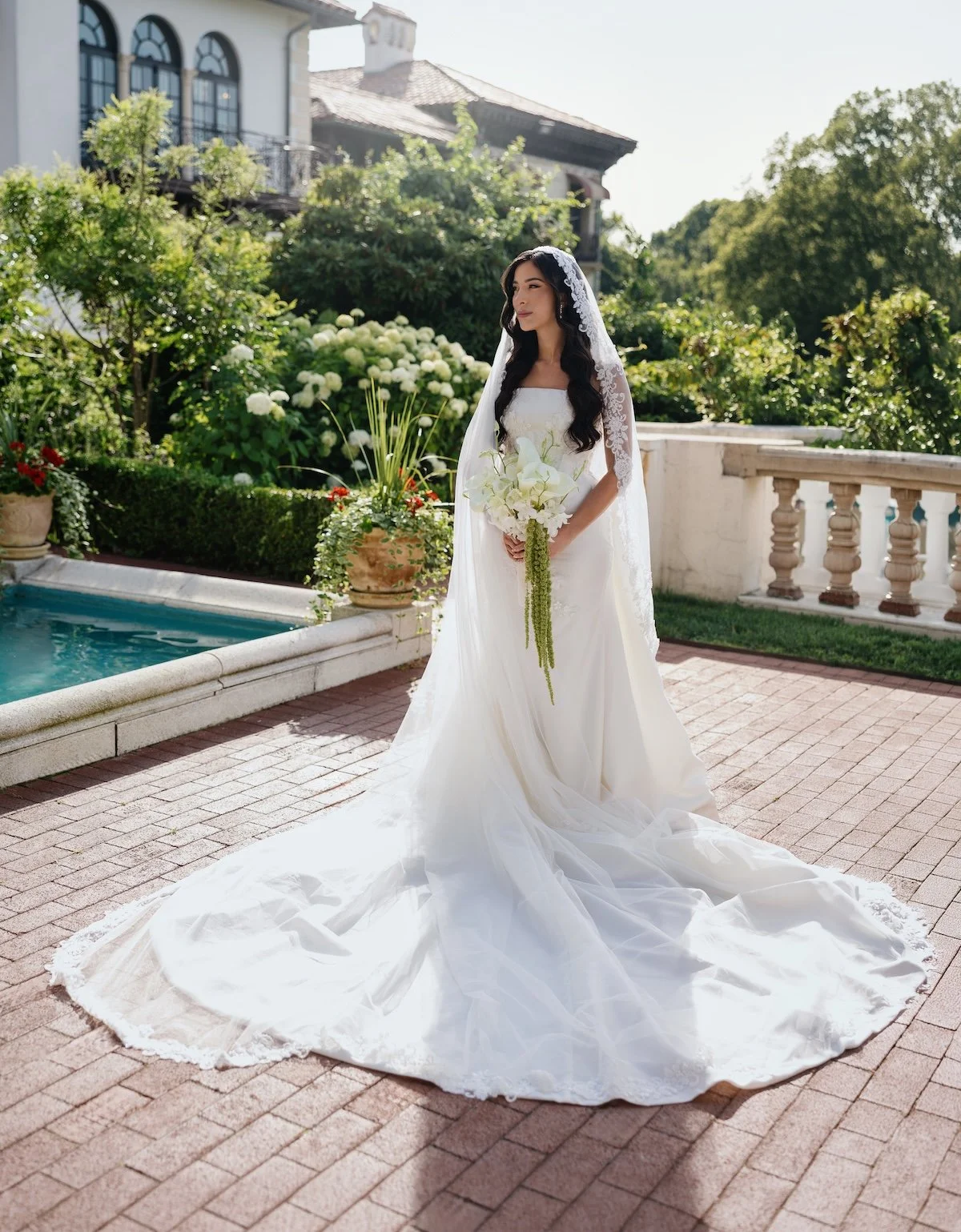 A bride in a white wedding dress holding a bouquet of white flowers, standing on a brick patio beside a small pool, with lush green plants and a large house in the background.