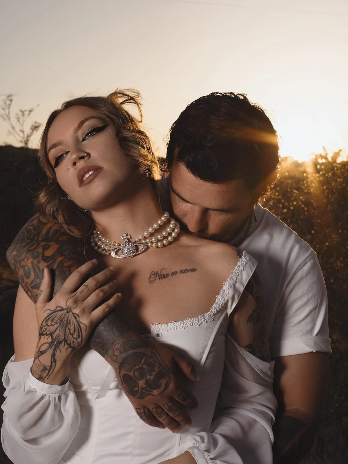 A bride with tattoos and a pearl necklace, and a man embracing her from behind during sunset.