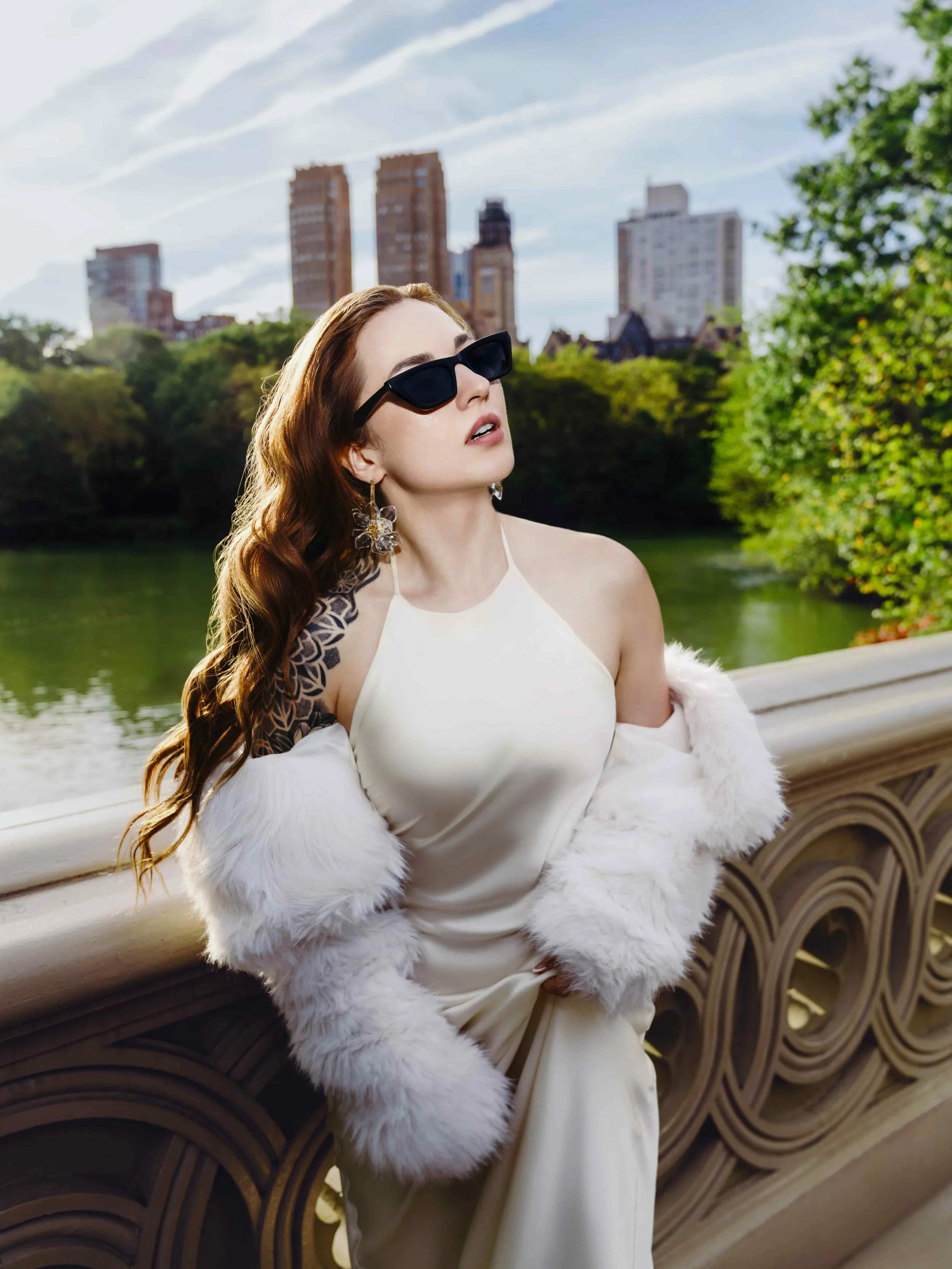 A woman with wavy auburn hair wearing black sunglasses, a white satin dress, and a white fur coat draped over her arms, standing on a bridge overlooking a river with city buildings and trees in the background.