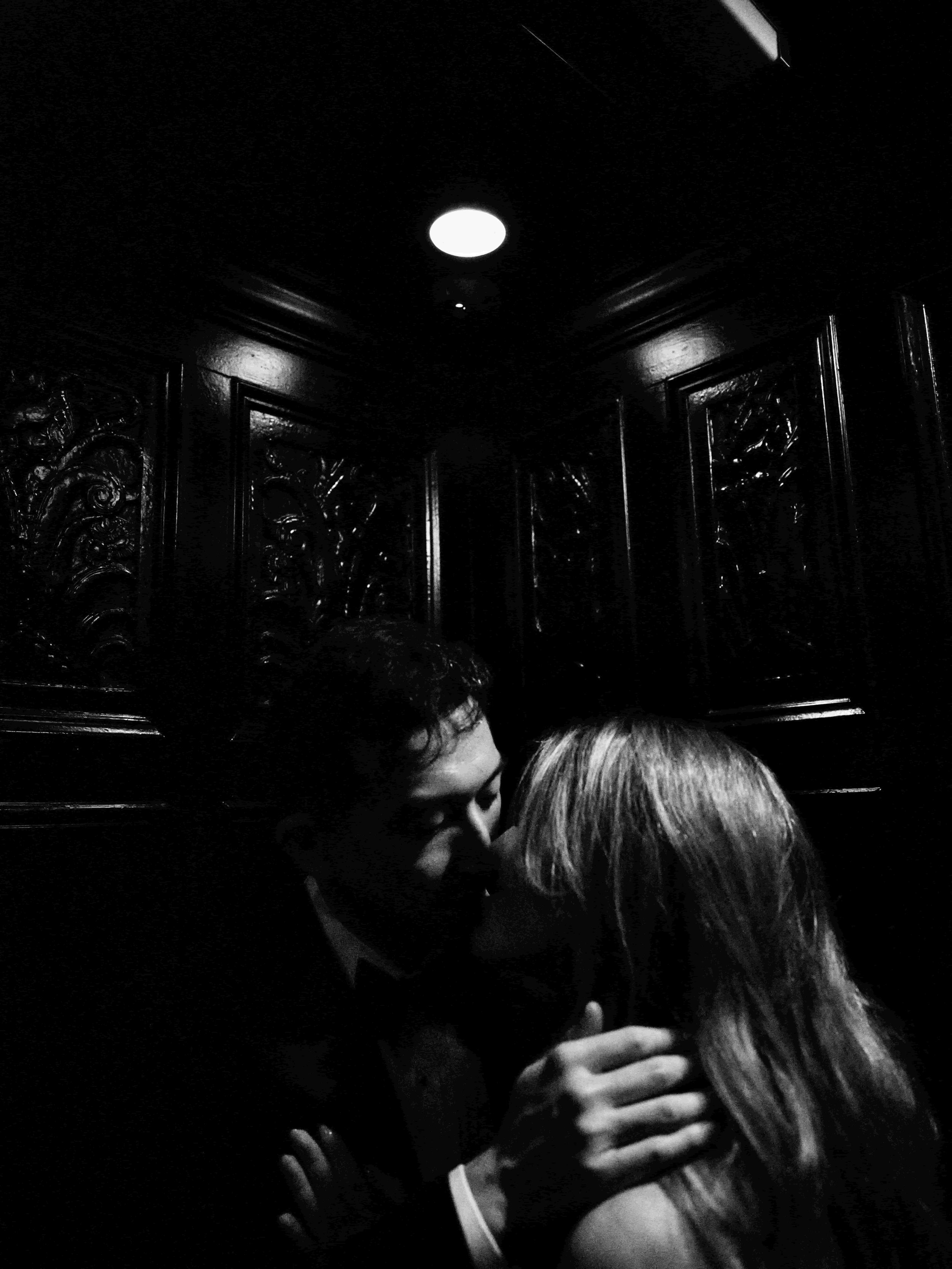 A black and white photo of a couple kissing in a dark, ornate room, with a bright ceiling light overhead.