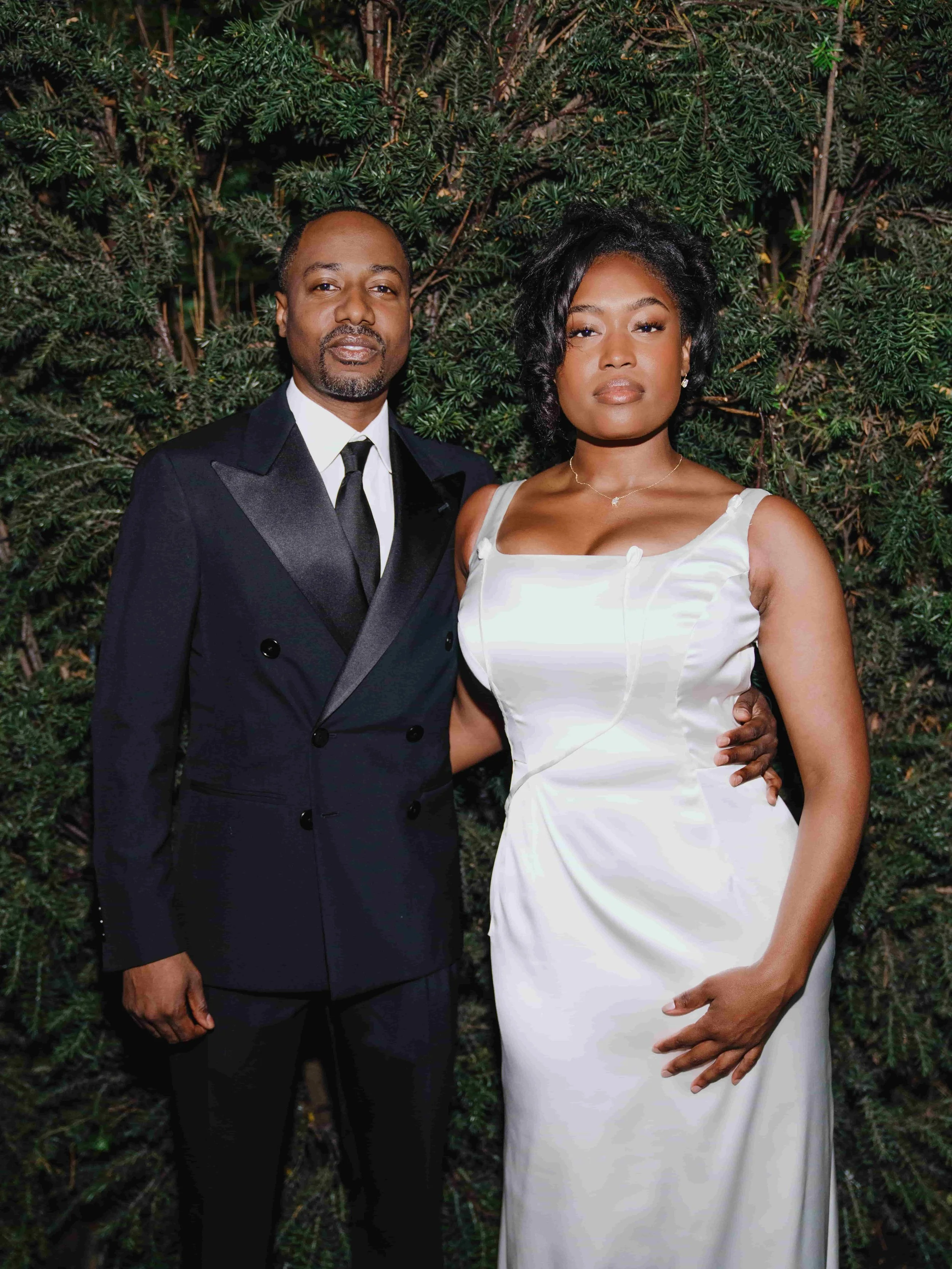 A groom in a black tuxedo and a bride in a white satin dress stand together outdoors in front of green foliage, posing for a photo.